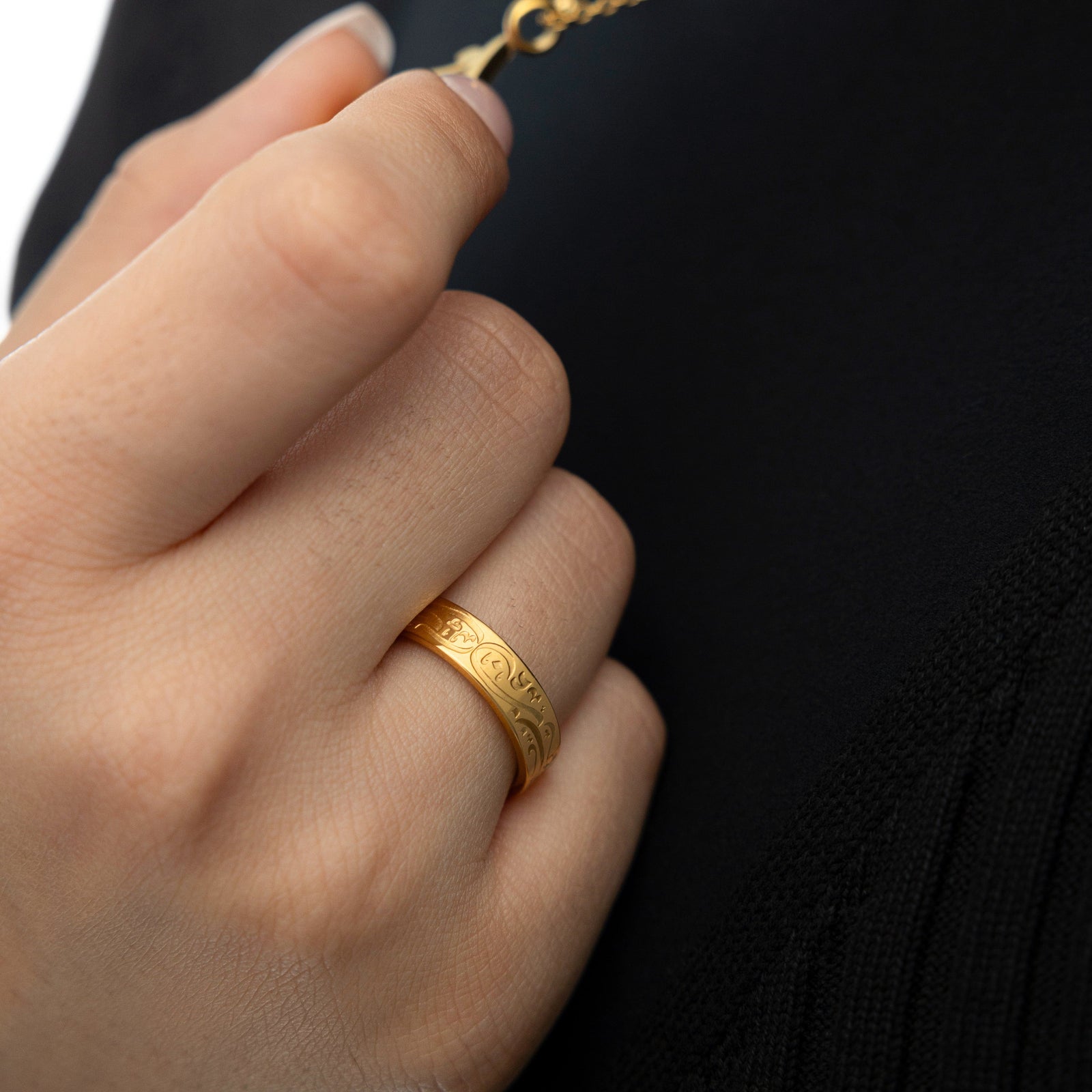 Close-up of a hand gently holding a necklace, wearing the gold “Be, and it is” ring with engraved Arabic text, styled with a black top.