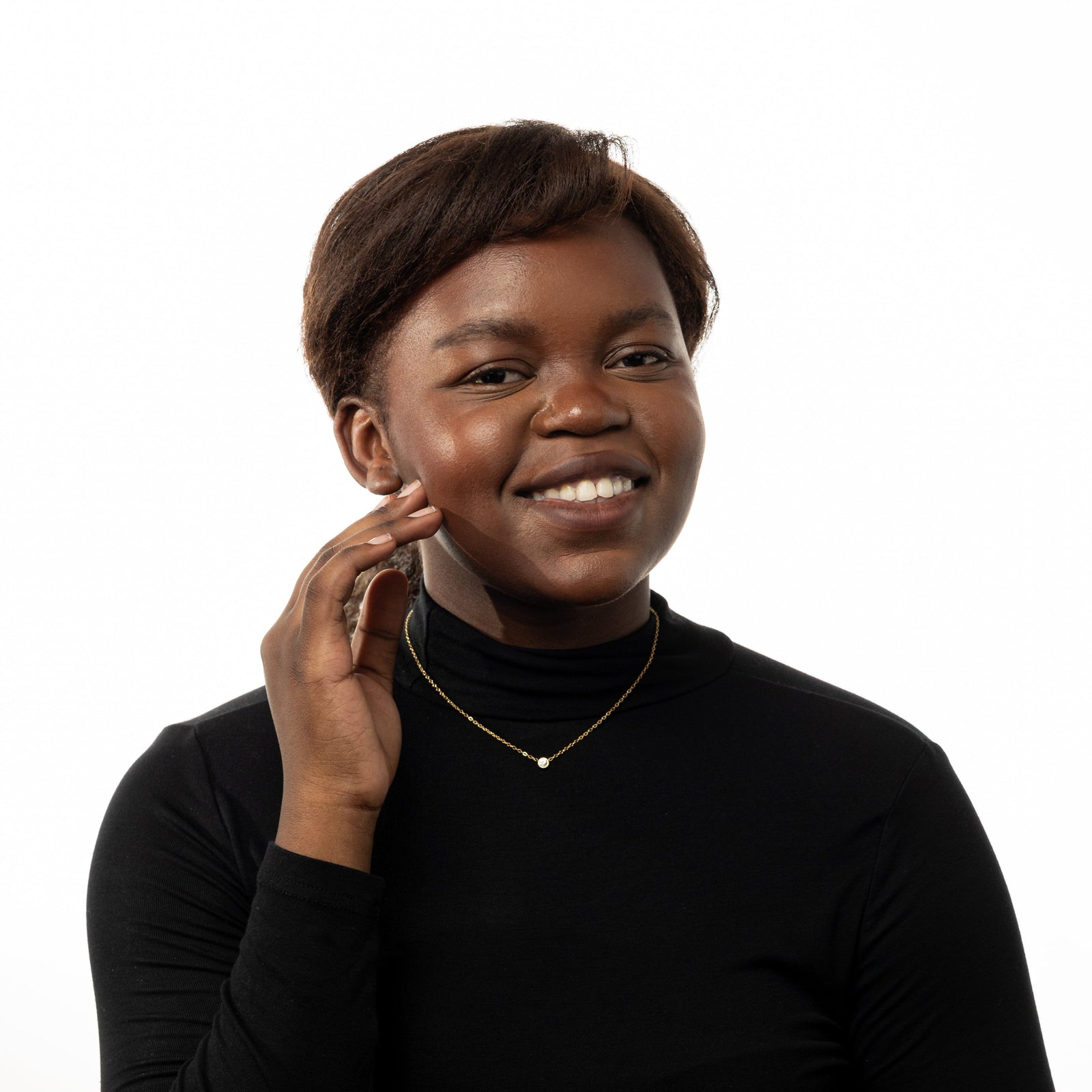 Photo of the model smiling to the camera while they gently touch their right cheek with their right hand. The model is wearing the gold Bezel Necklace.