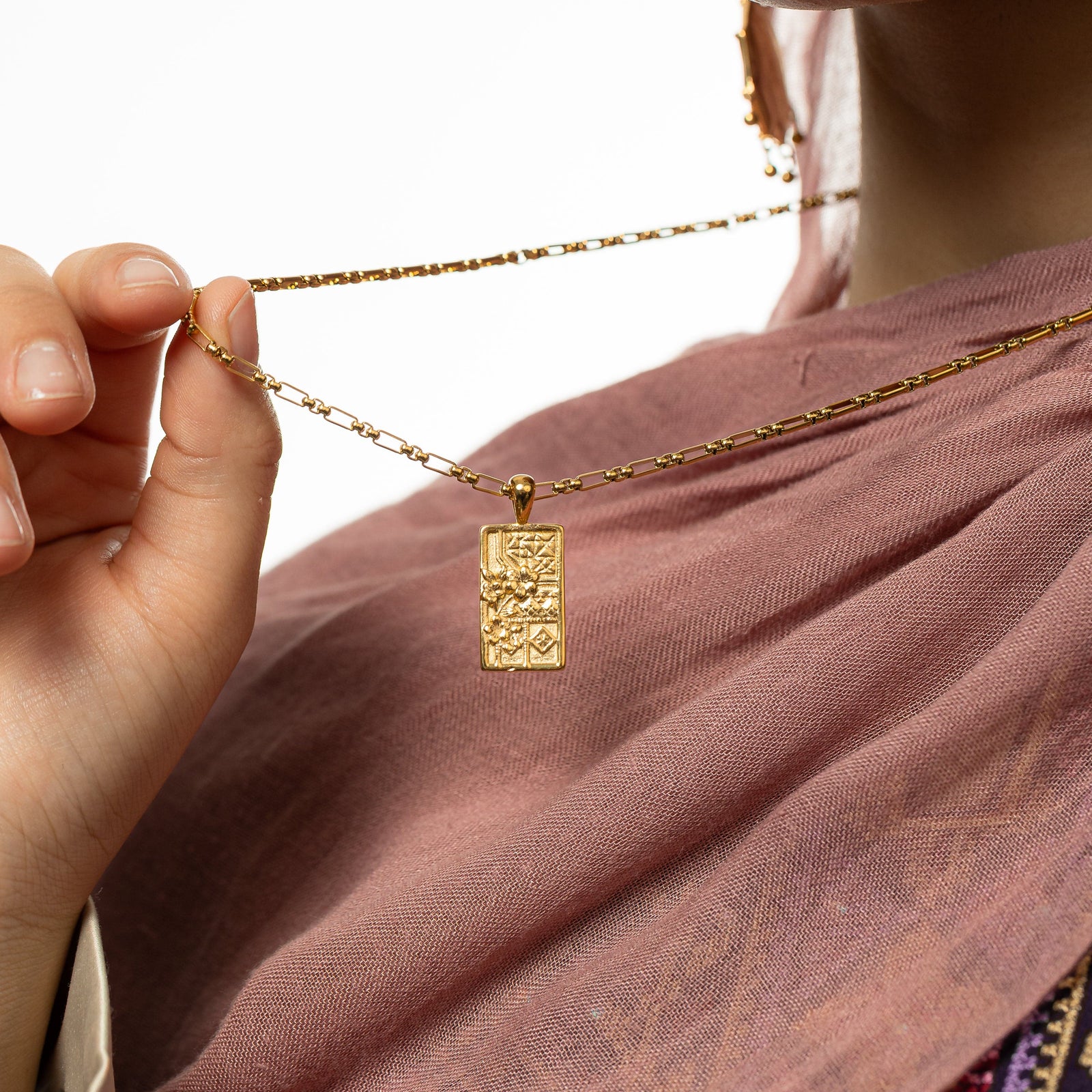 Close up photo of model holding the gold chain of the necklace to show the design of the rectangular pendant.