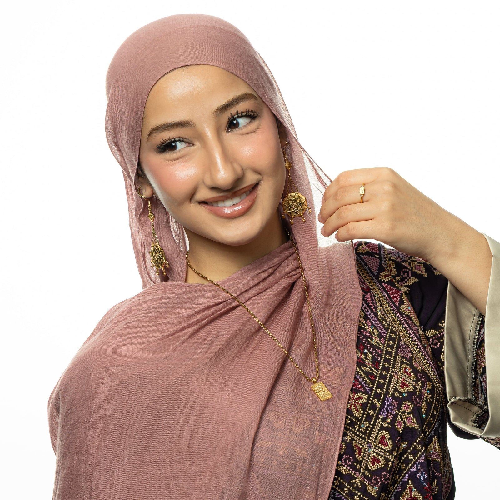 Photo of the model looking and smiling away from the camera wearing the gold Damascus Jasmine Mosaic Necklace, gold Ummi | Mother Coin Earrings, and gold We are Strong ring.