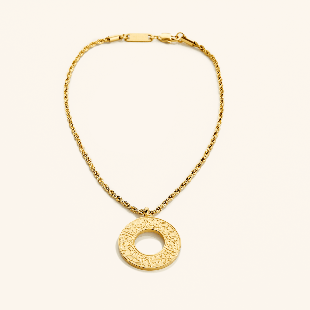 Gold car hanger with a circular pendant engraved in Arabic calligraphy reading “With Hardship Comes Ease,” displayed against a cream background.