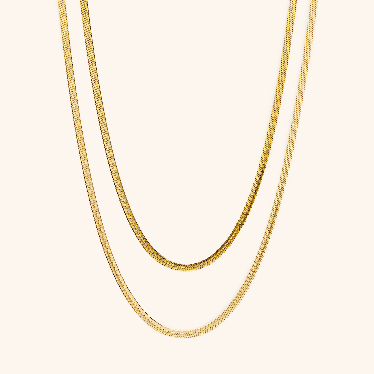 Two gold herringbone chains laid flat against a cream background.