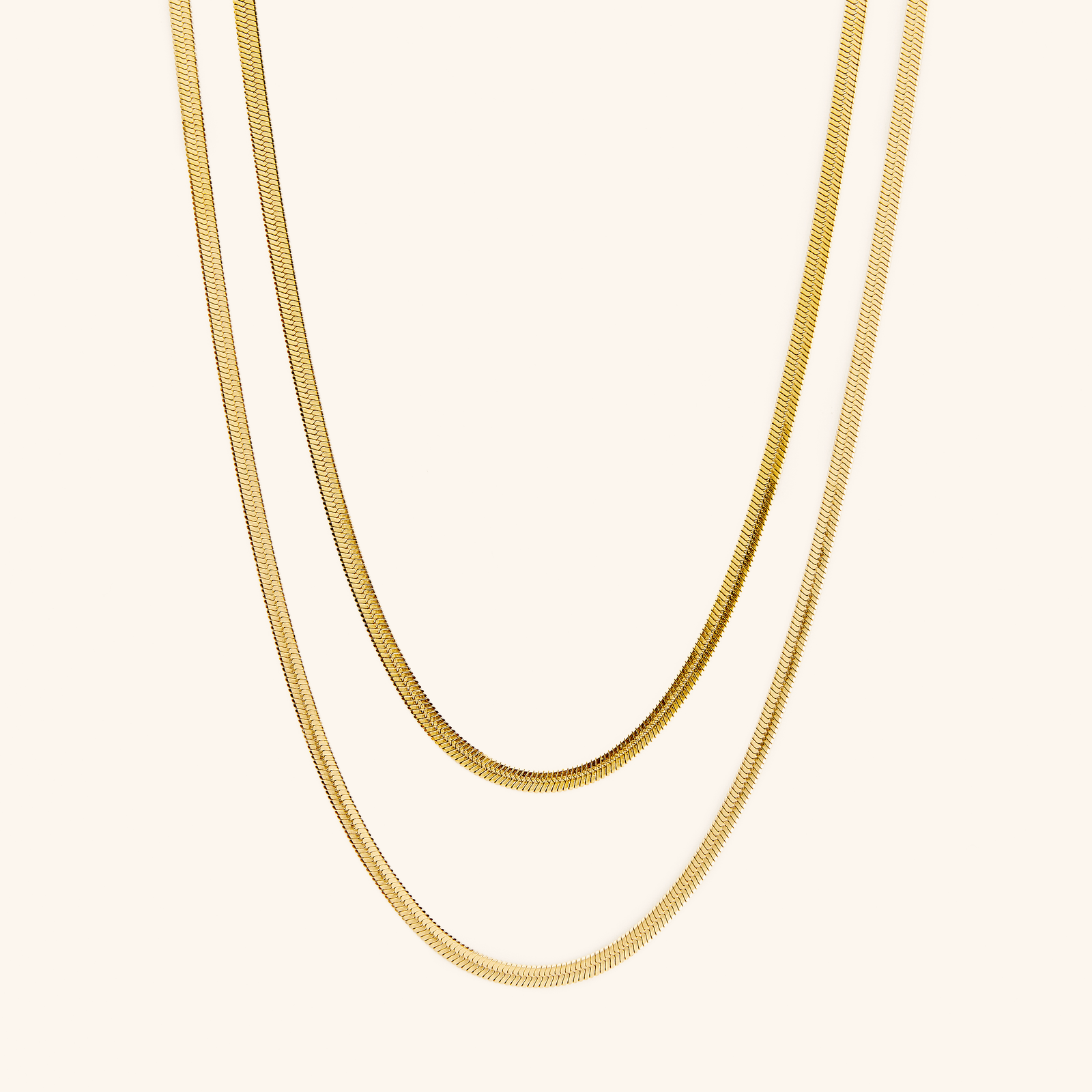 Two gold herringbone chains laid flat against a cream background.