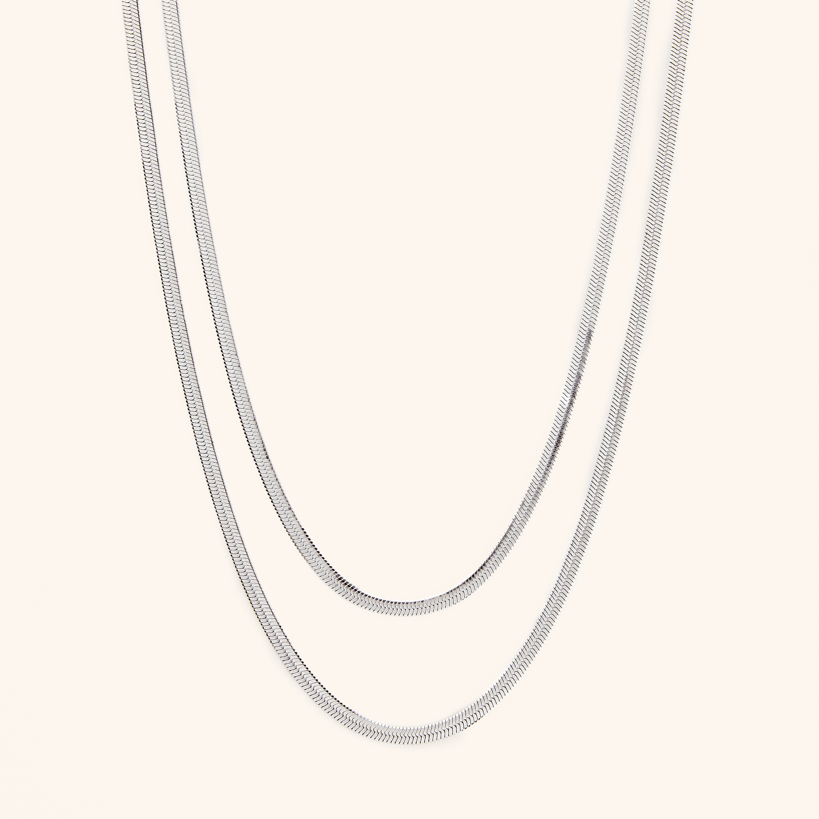  Two silver herringbone chains laid flat against a cream background.