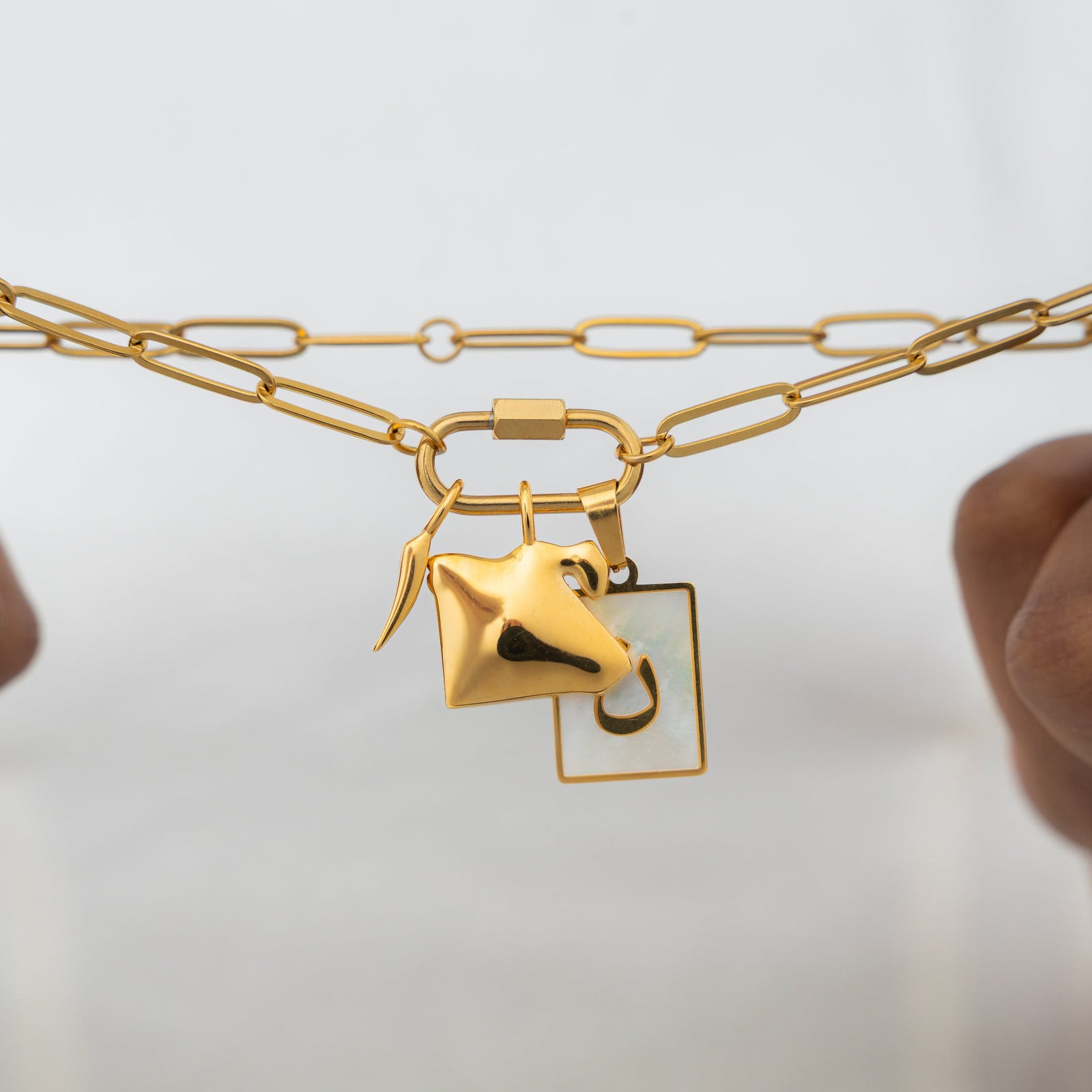 A close-up of the gold-plated Charm Keeper Necklace held in a model's hands, highlighting the carabiner clasp and attached charms.