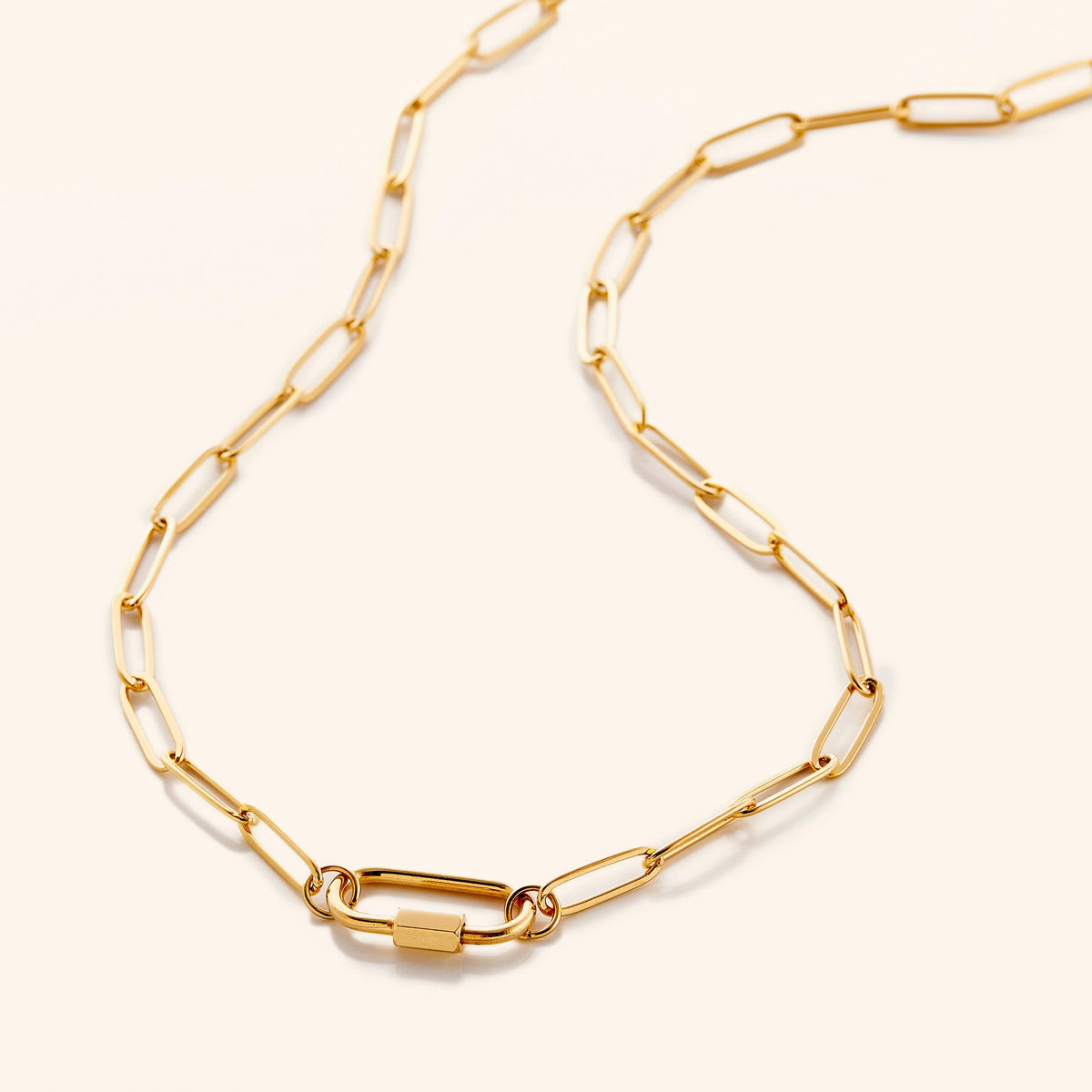 A close-up of a gold-plated Charm Keeper Necklace featuring a sleek carabiner clasp, with a rectangular charm and a square charm attached, resting on a model's open palm.