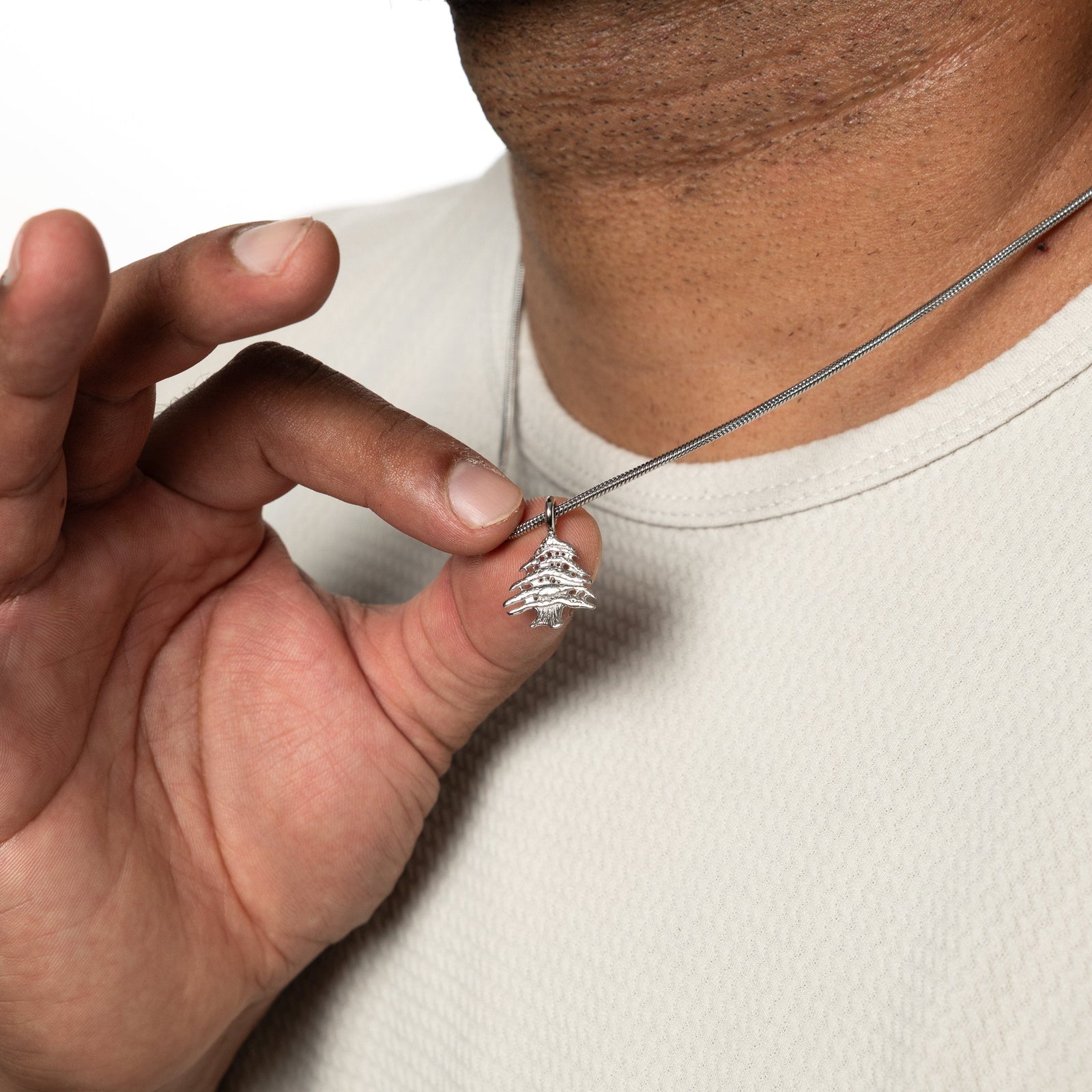 Man holding up a silver Cedar of Lebanon necklace, worn over a white textured shirt.