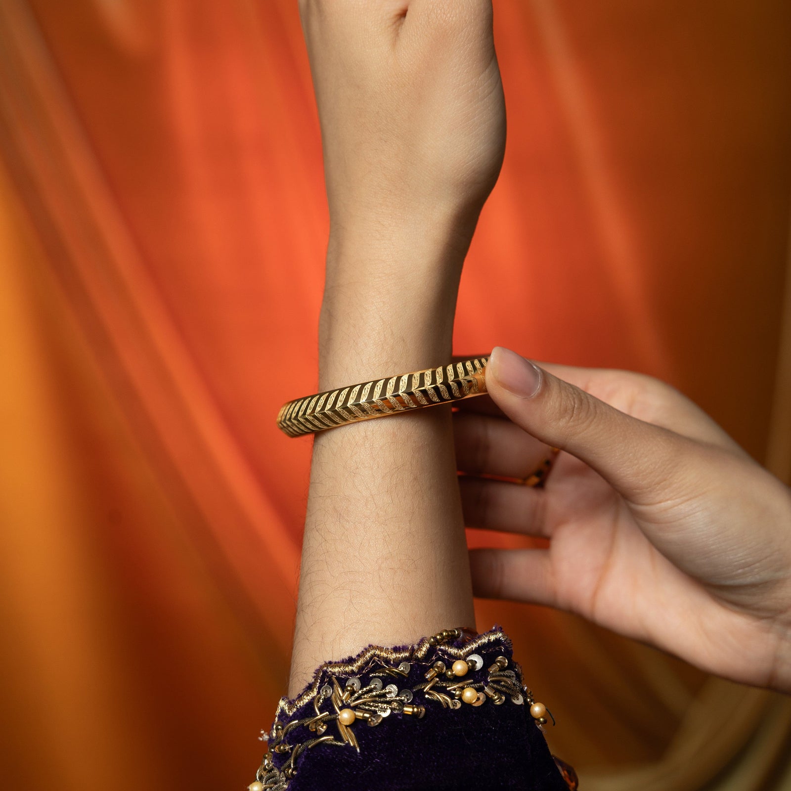 A person's arm is shown with a gold bangle featuring a repeating chevron or zigzag pattern being placed on their wrist by another hand. The person wearing the bangle is wearing a dark purple sleeve with gold embroidery and embellishments. The background is a gradient of orange and yellow.
