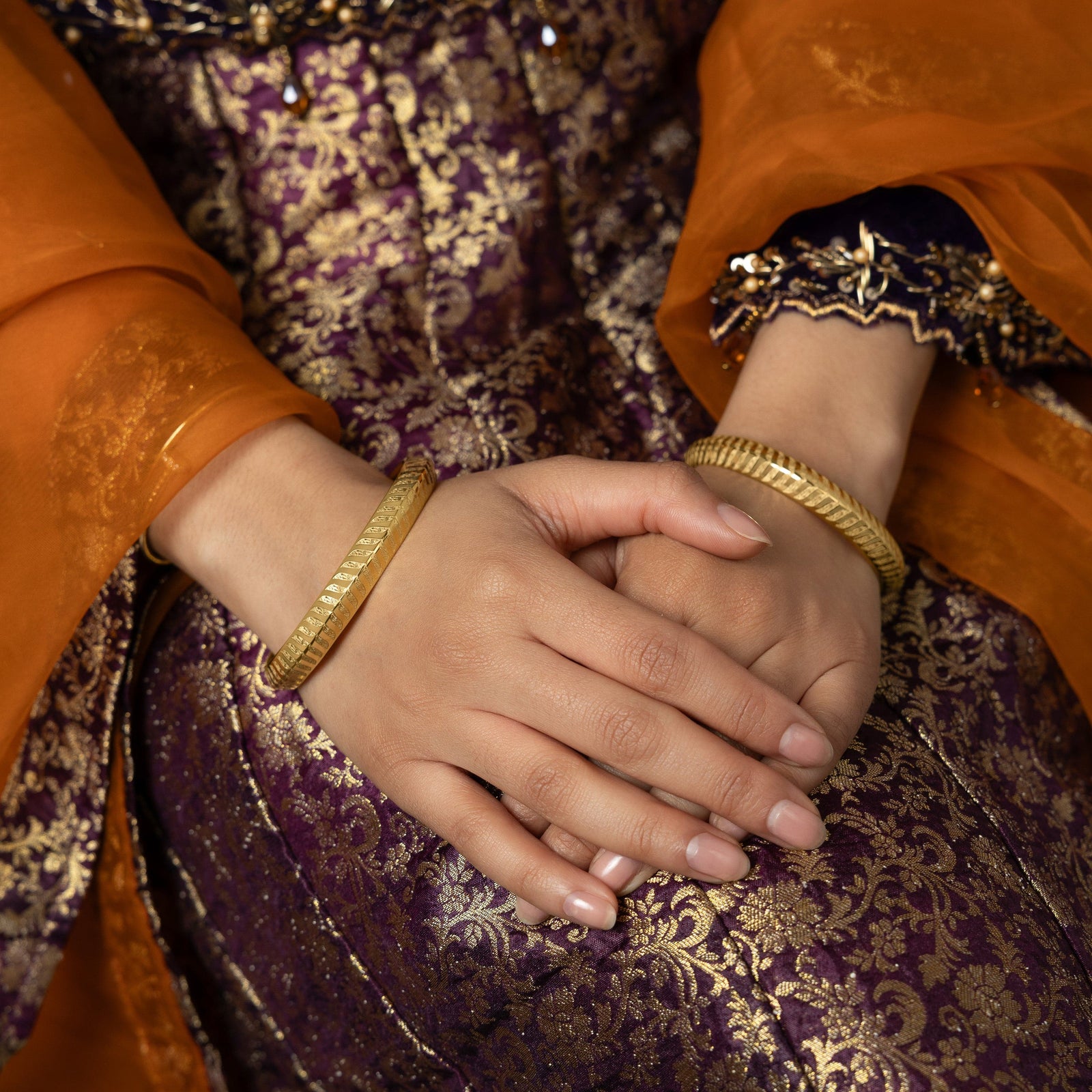 A person's hands are clasped together, each wrist adorned with a gold bangle featuring a repeating chevron or zigzag pattern. The person is wearing a richly textured purple and gold garment with sheer orange fabric layered over the sleeves.