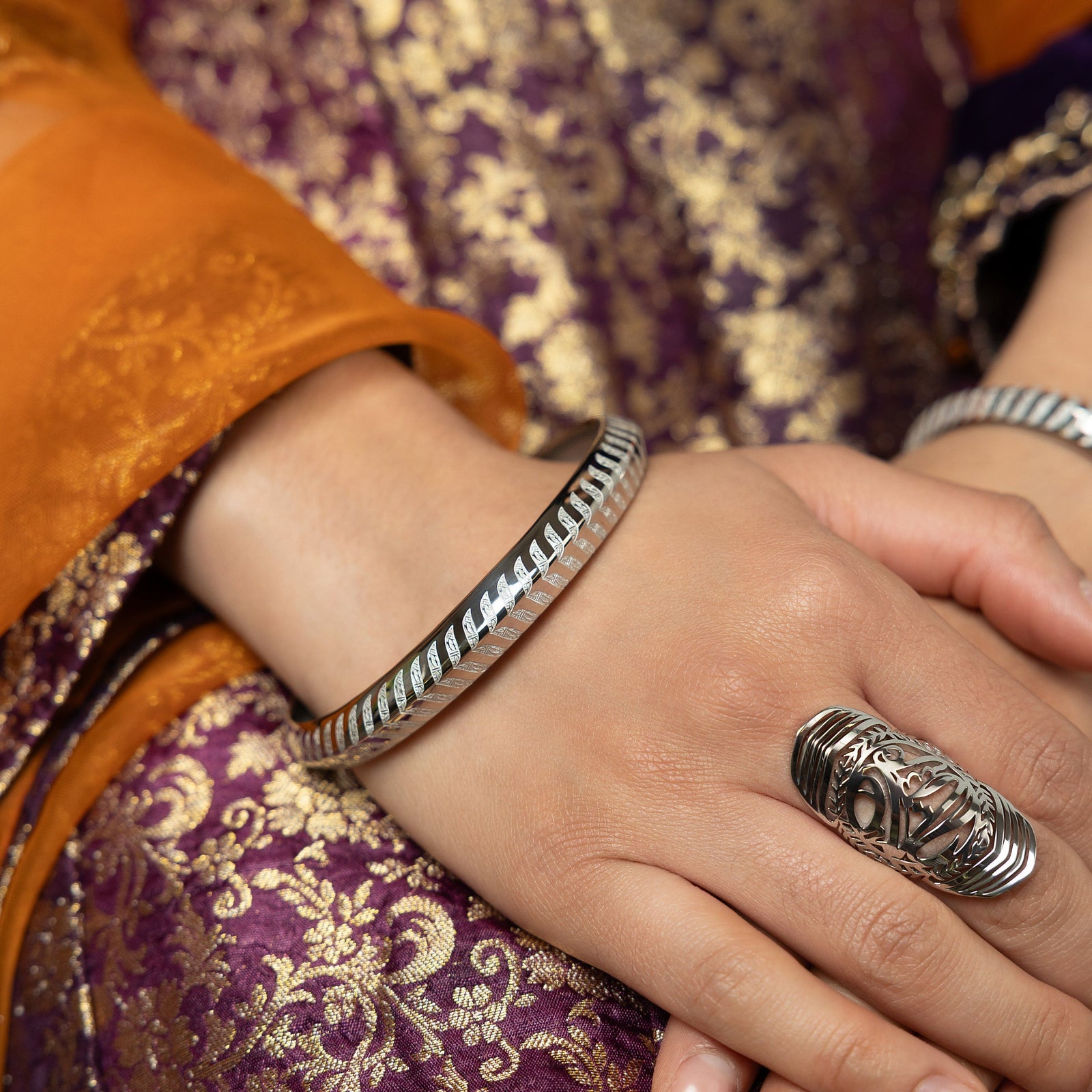 A person wearing a patterned purple and gold garment with an orange sleeve has a silver bangle with a chevron pattern on their wrist and an intricate silver cut-out ring on their middle finger.