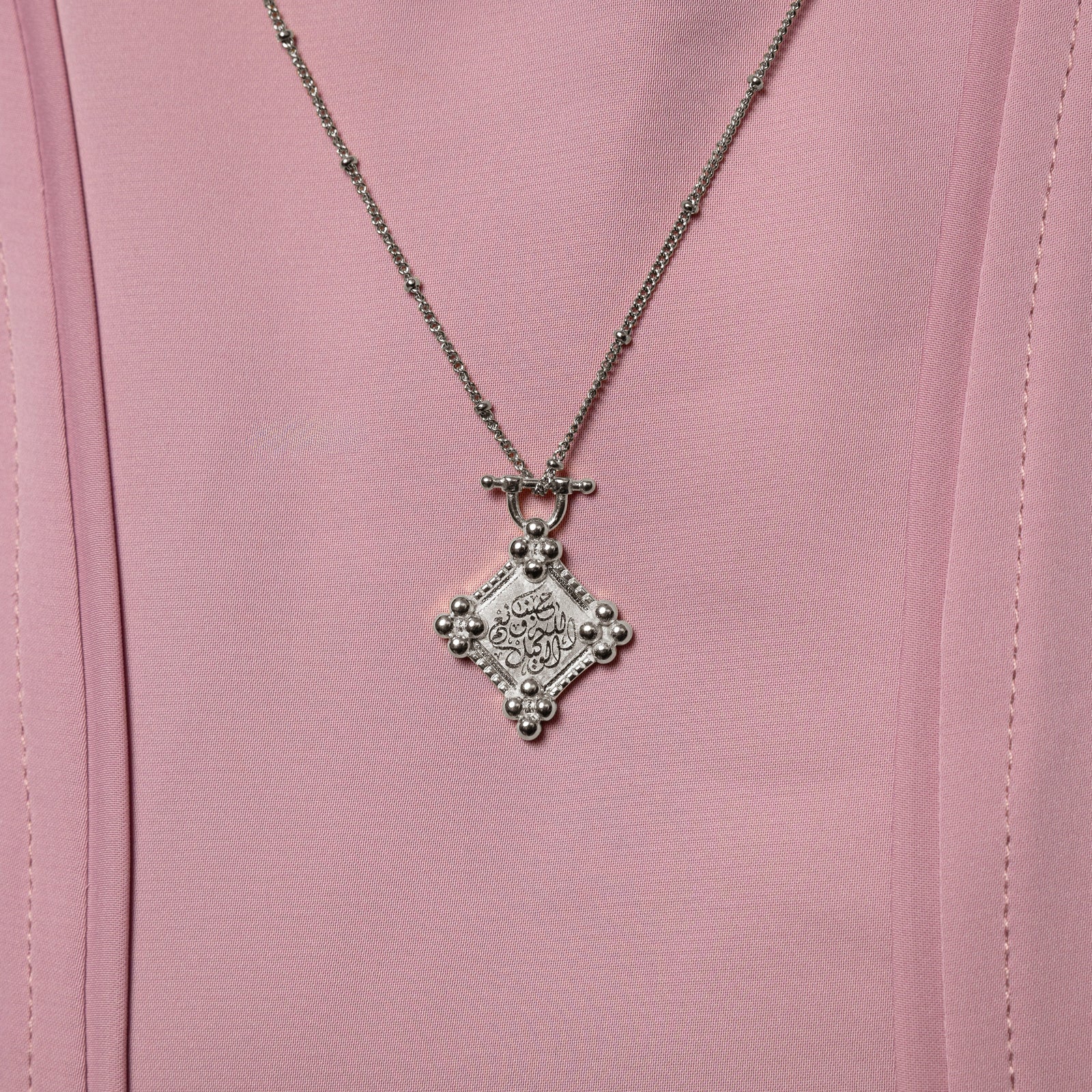 Close up of the silver "Allah is sufficient for us" Plaque Necklace on a pink background.
