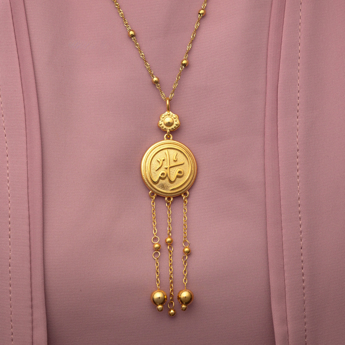 Close up of the gold Coin Mama Necklace pendant on a pink fabric background.