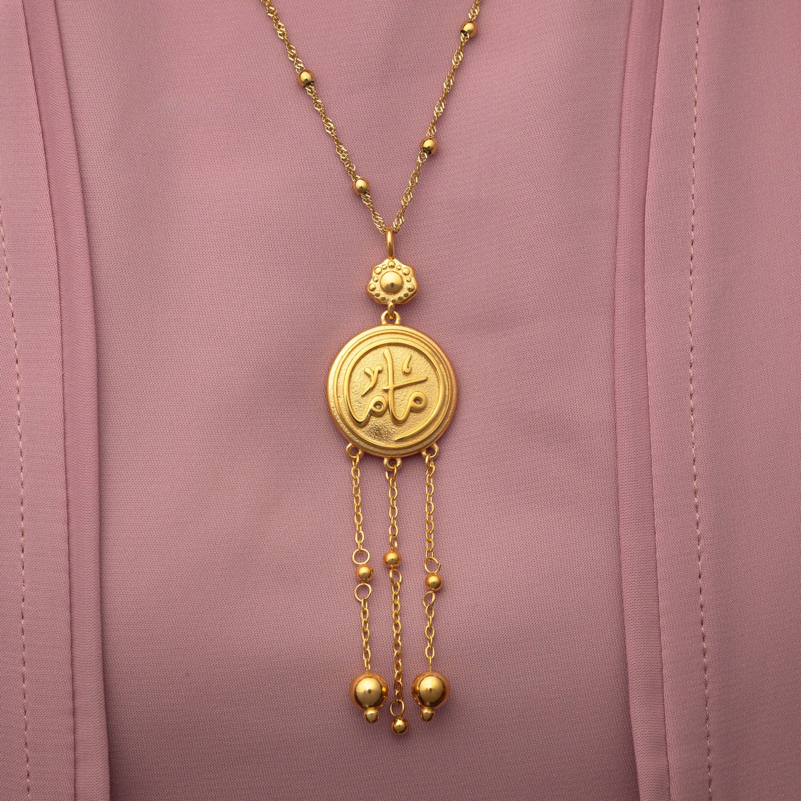 Close up of the gold Coin Mama Necklace pendant on a pink fabric background.