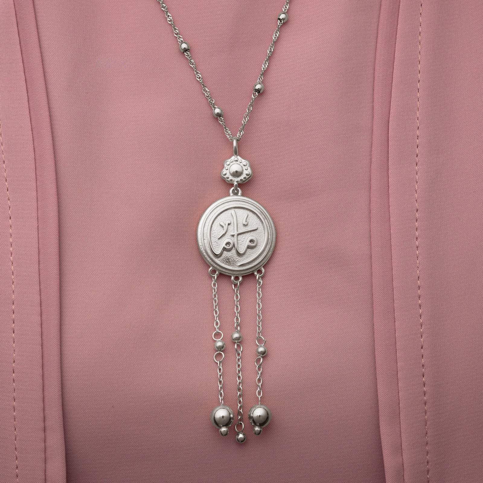 Close up of the silver Coin Mama Necklace pendant on a pink fabric background.