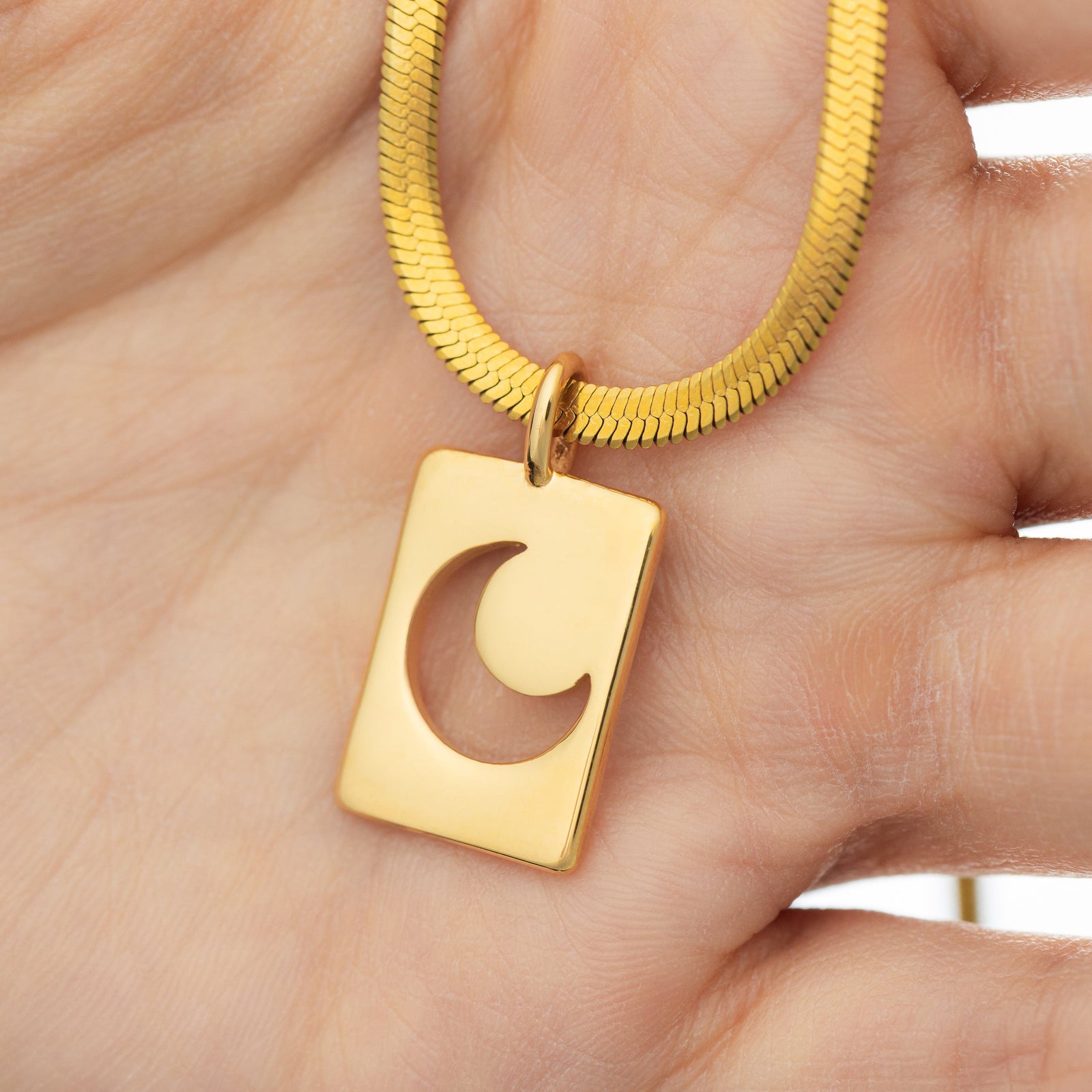 Gold crescent charm hanging from a thick snake chain, shown up close against fingers for size reference and texture.
