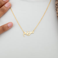 A delicate gold-plated chain necklace featuring a pendant in the shape of stylized Arabic script. The necklace is displayed against a white background. The Arabic script appears to spell out a name.