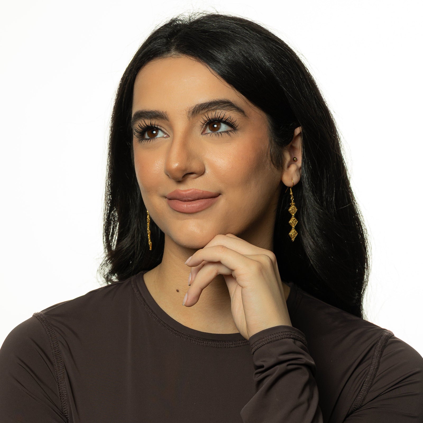 Photo of the model smiling and touching their chin with their left hand knuckles while wearing the gold Draping Thikr Earrings.