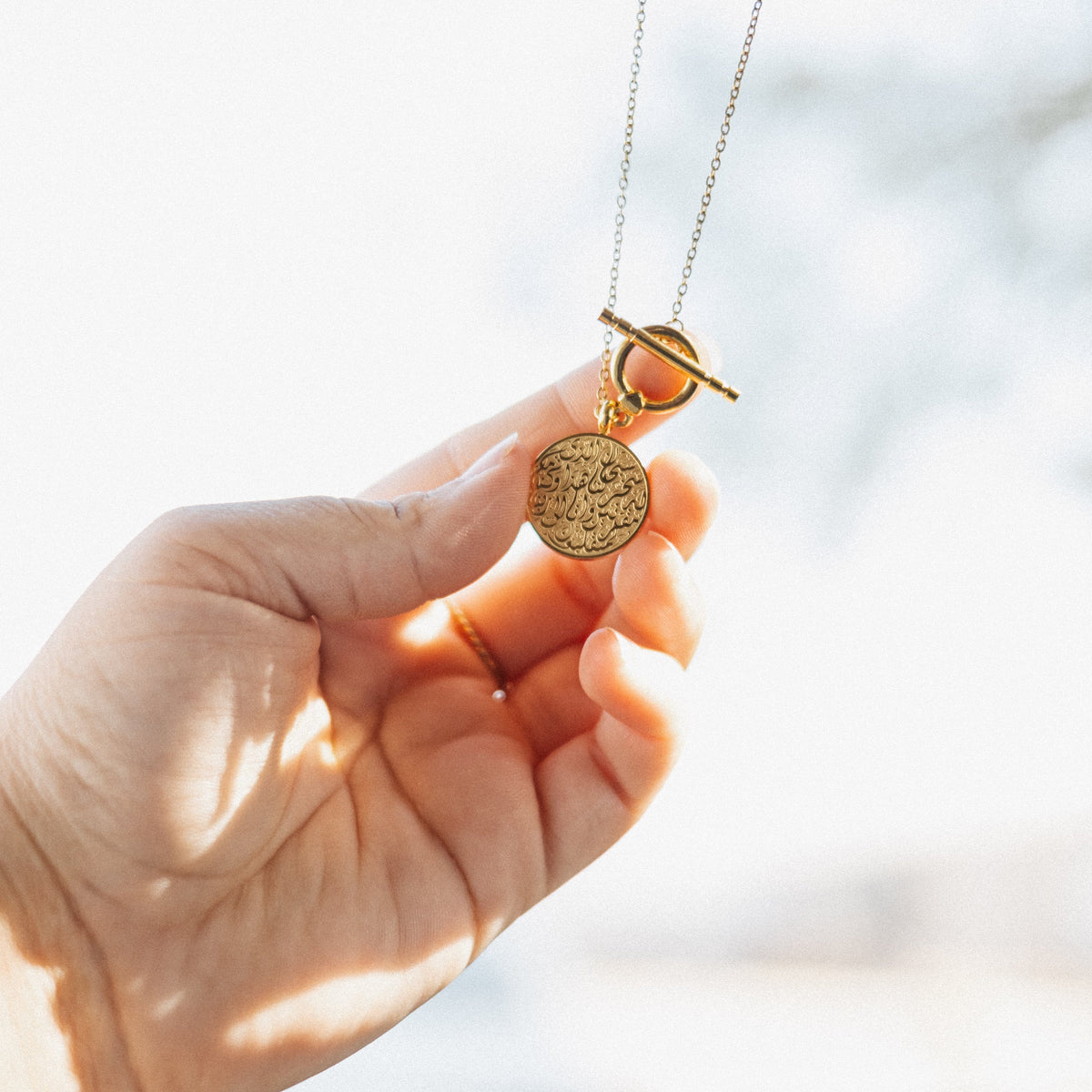 Hand holding the gold Duaa Al-Safar pendant up, the Arabic calligraphy details visible on the medallion.