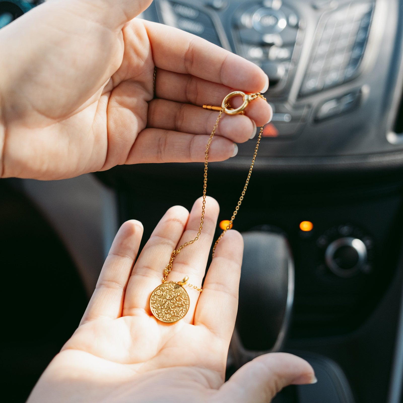 Hands holding the gold Duaa Al-Safar car hanger in front of a car dashboard, pendant reflecting light with engraved Arabic calligraphy clearly seen.