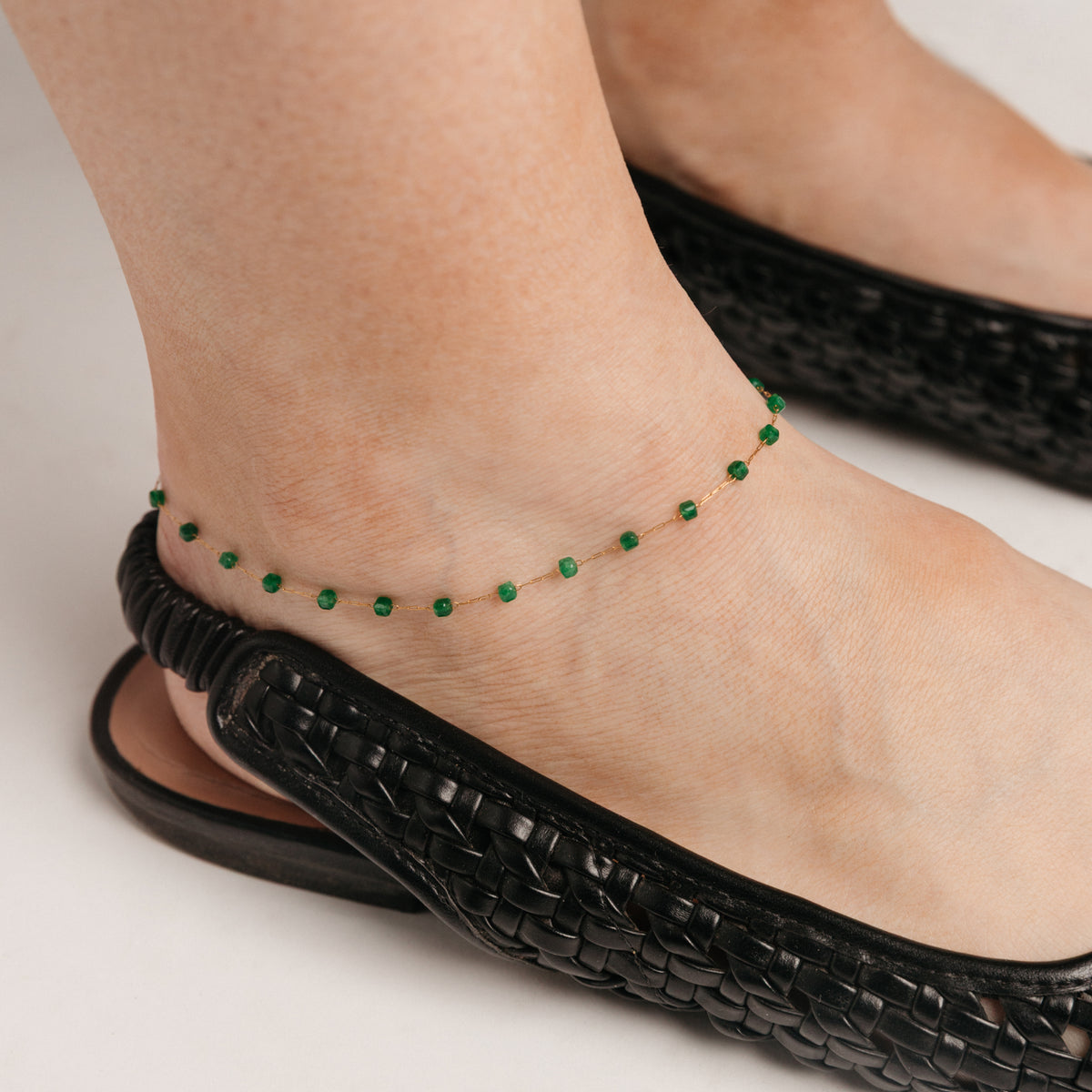 Close-up of an ankle wearing an 18k gold-plated emerald-beaded chain anklet and a black woven shoe