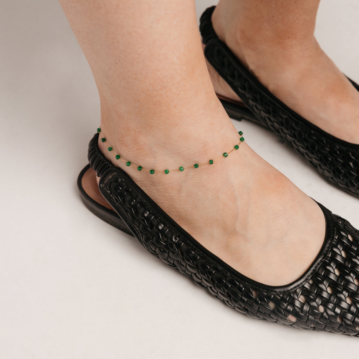 Close-up of an ankle wearing an 18k gold-plated emerald-beaded chain anklet and a black woven shoe