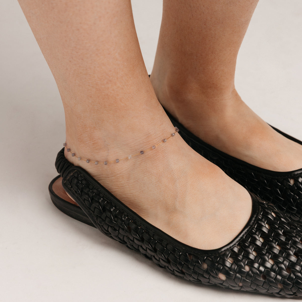 Close-up view of a person's ankle wearing the Essential Iolite Micro Anklet, featuring a delicate 18k gold-plated chain accented with faceted violet-blue beads.