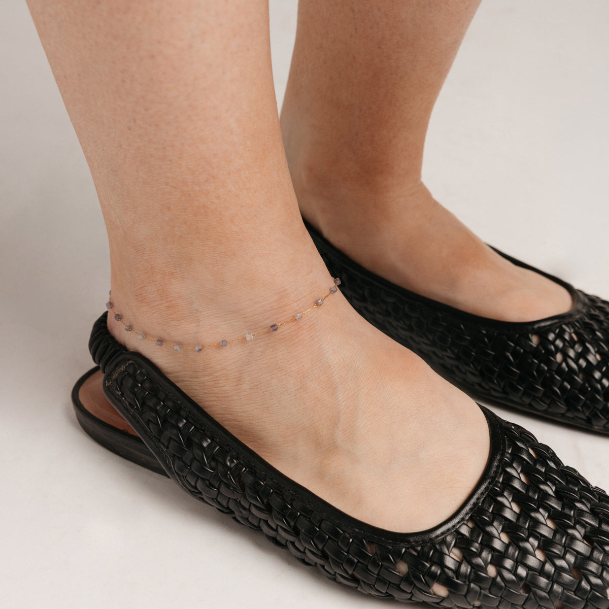 Close-up view of a person's ankle wearing the Essential Iolite Micro Anklet, featuring a delicate 18k gold-plated chain accented with faceted violet-blue beads.