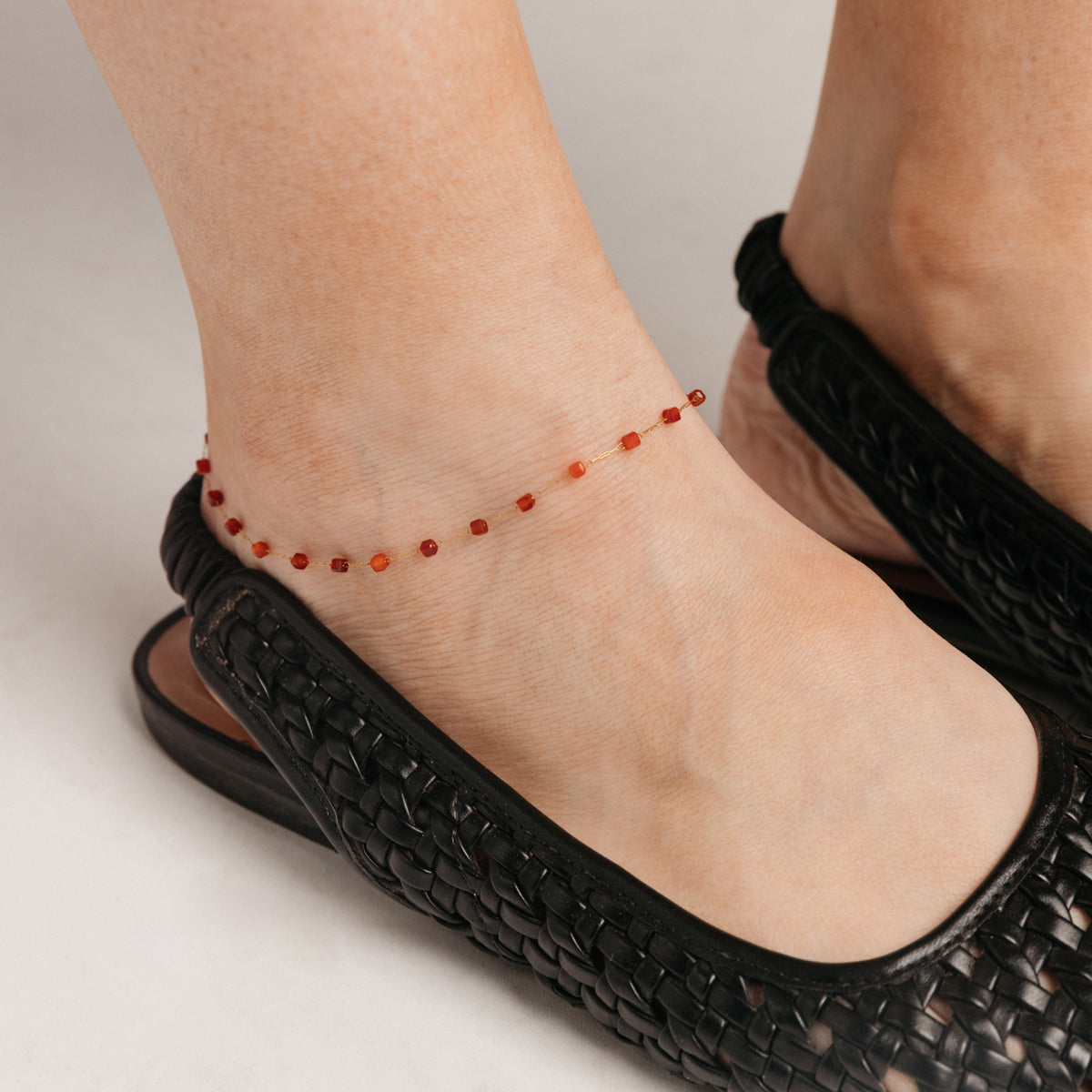 Close-up view of a person's ankle wearing the Essential Red Agate Micro Anklet, featuring a delicate 18k gold-plated chain accented with red agate beads, styled alongside black woven footwear.