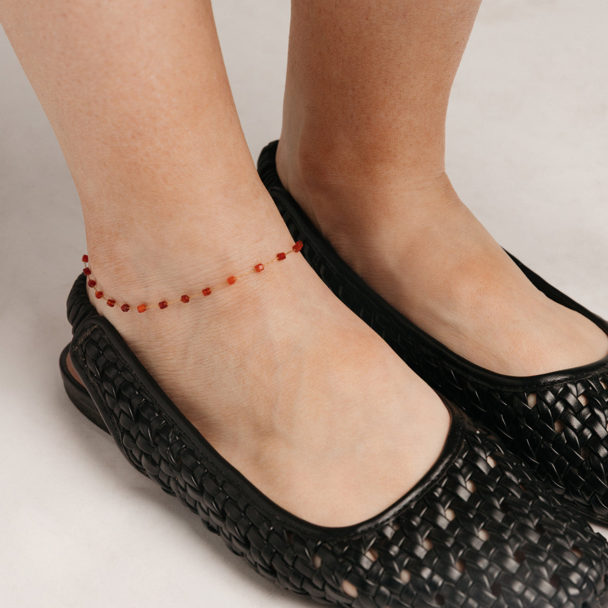 Close-up view of a person's ankle wearing the Essential Red Agate Micro Anklet, featuring a delicate 18k gold-plated chain accented with red agate beads, styled alongside black woven footwear.