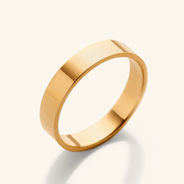 A polished, flat-edge 18k gold-plated band ring angled diagonally against a light cream background.