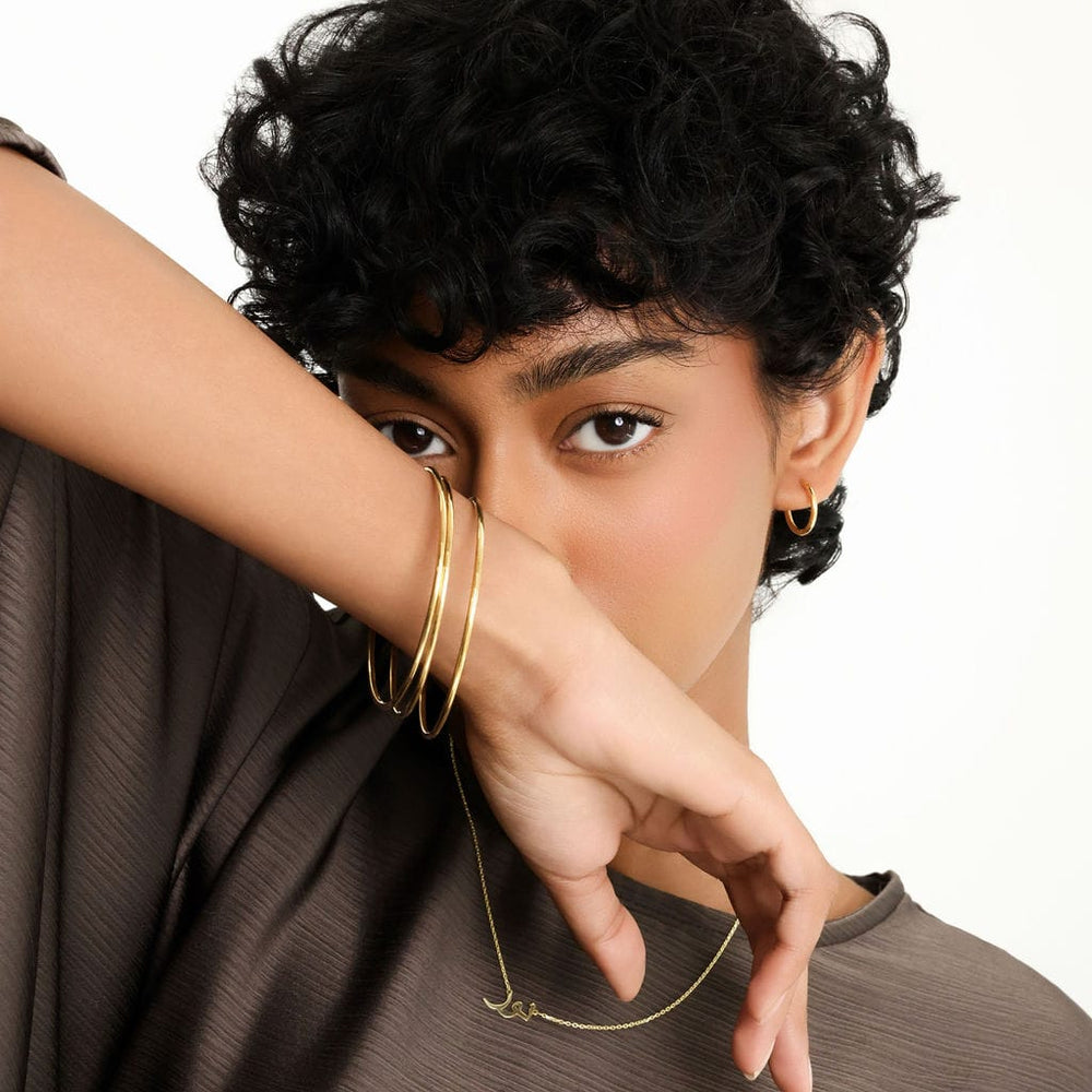 Photo of the model with their arms up covering the bottom half of her face. The model is wearing three pieces of gold Essential Thin Bangle on their right hand.