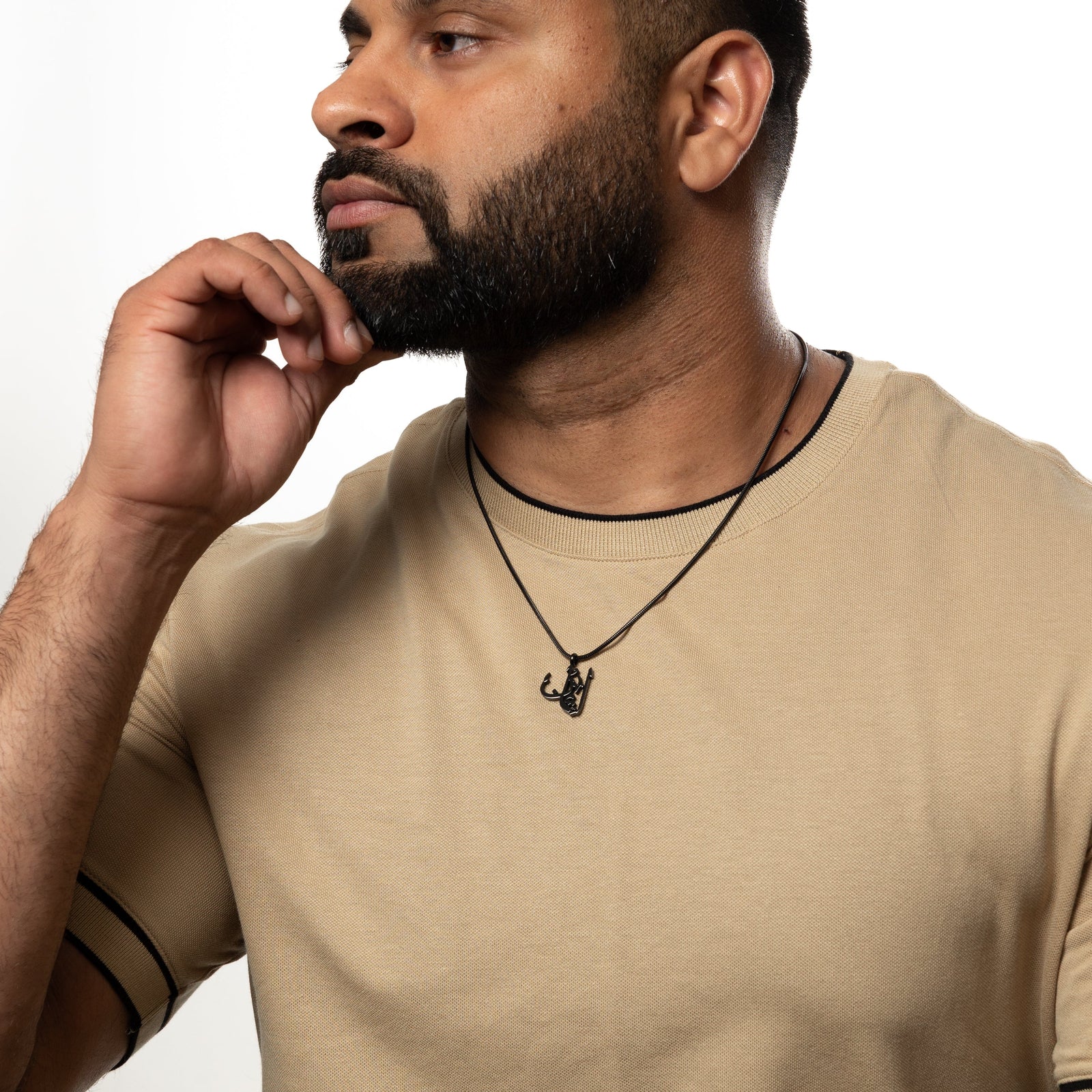 Man wearing a black 'Faith' calligraphy necklace with a beige shirt, resting hand on chin.