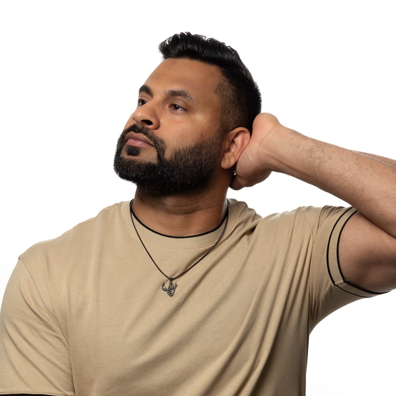 Man wearing a black 'Faith' calligraphy necklace over a beige shirt, hand behind head.