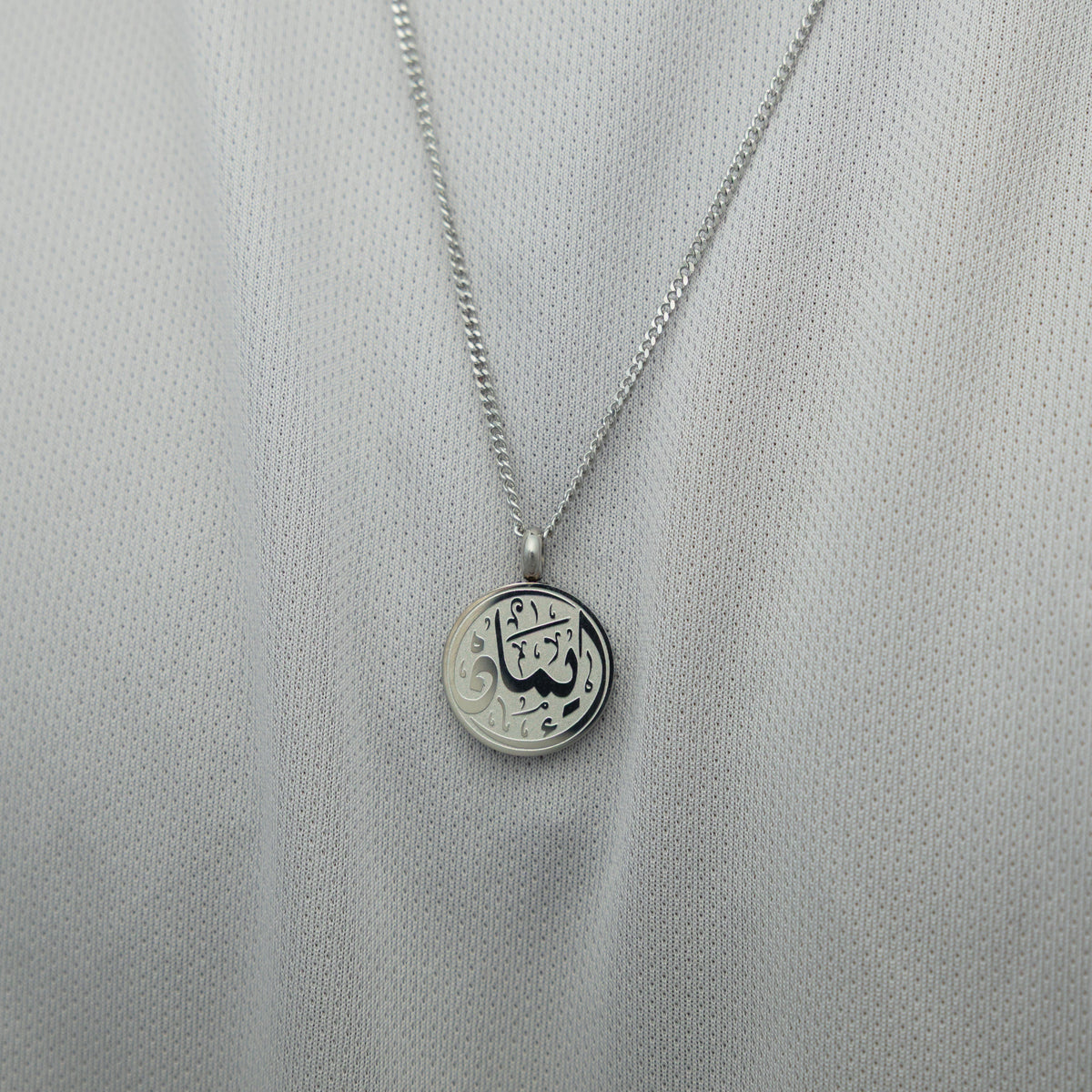 Silver faith token necklace over a white textured shirt, showing the engraved Arabic script “Iman”.