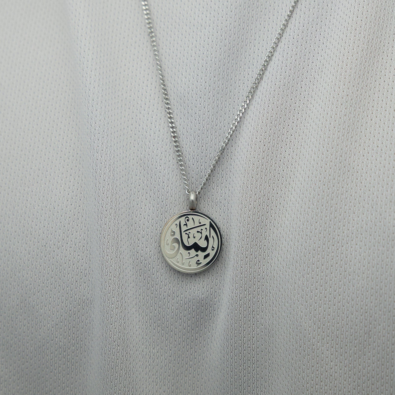 Silver faith token necklace over a white textured shirt, showing the engraved Arabic script “Iman”.
