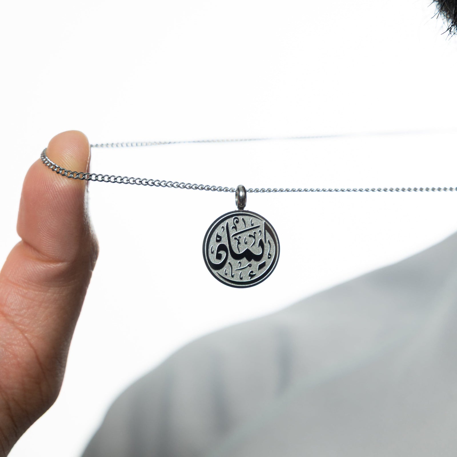 Man holding up a silver Faith Token necklace by the chain, against a light background.