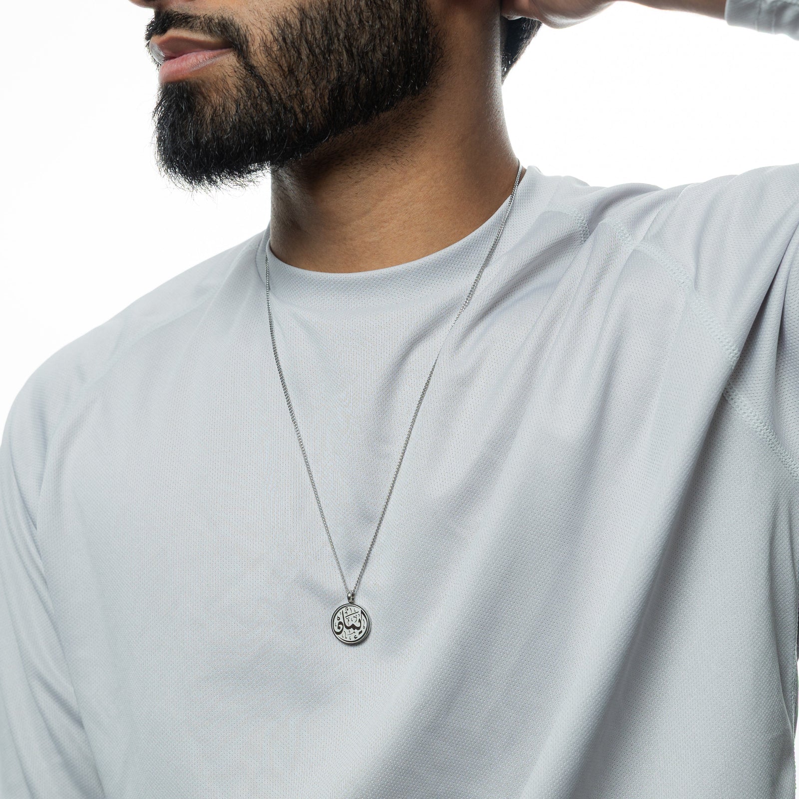 A silver Faith Token necklace worn over a white shirt, with the pendant centered on the chest, the man's hand resting behind his head.
