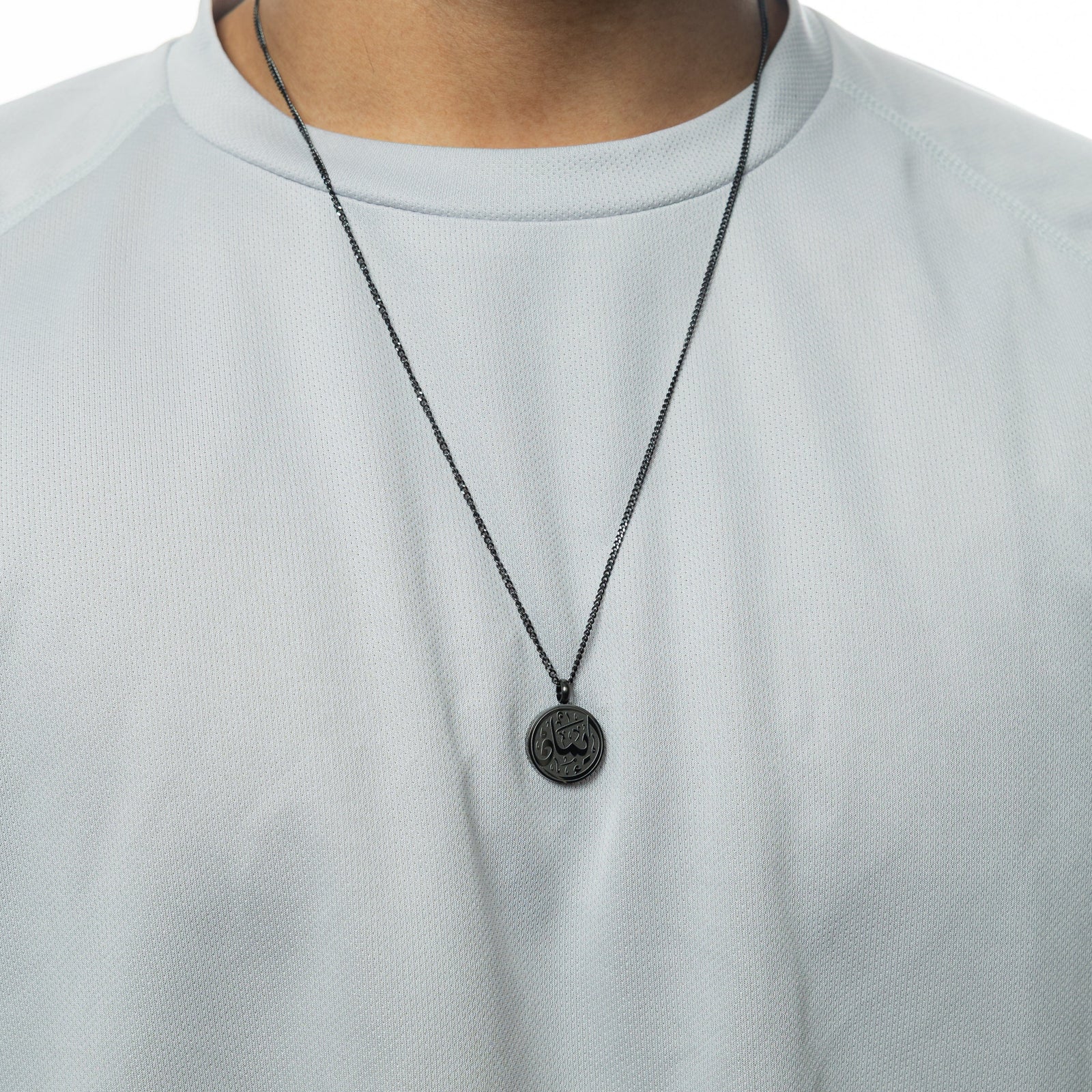 A black Faith Token necklace worn over a white textured shirt, pendant centered on the chest.