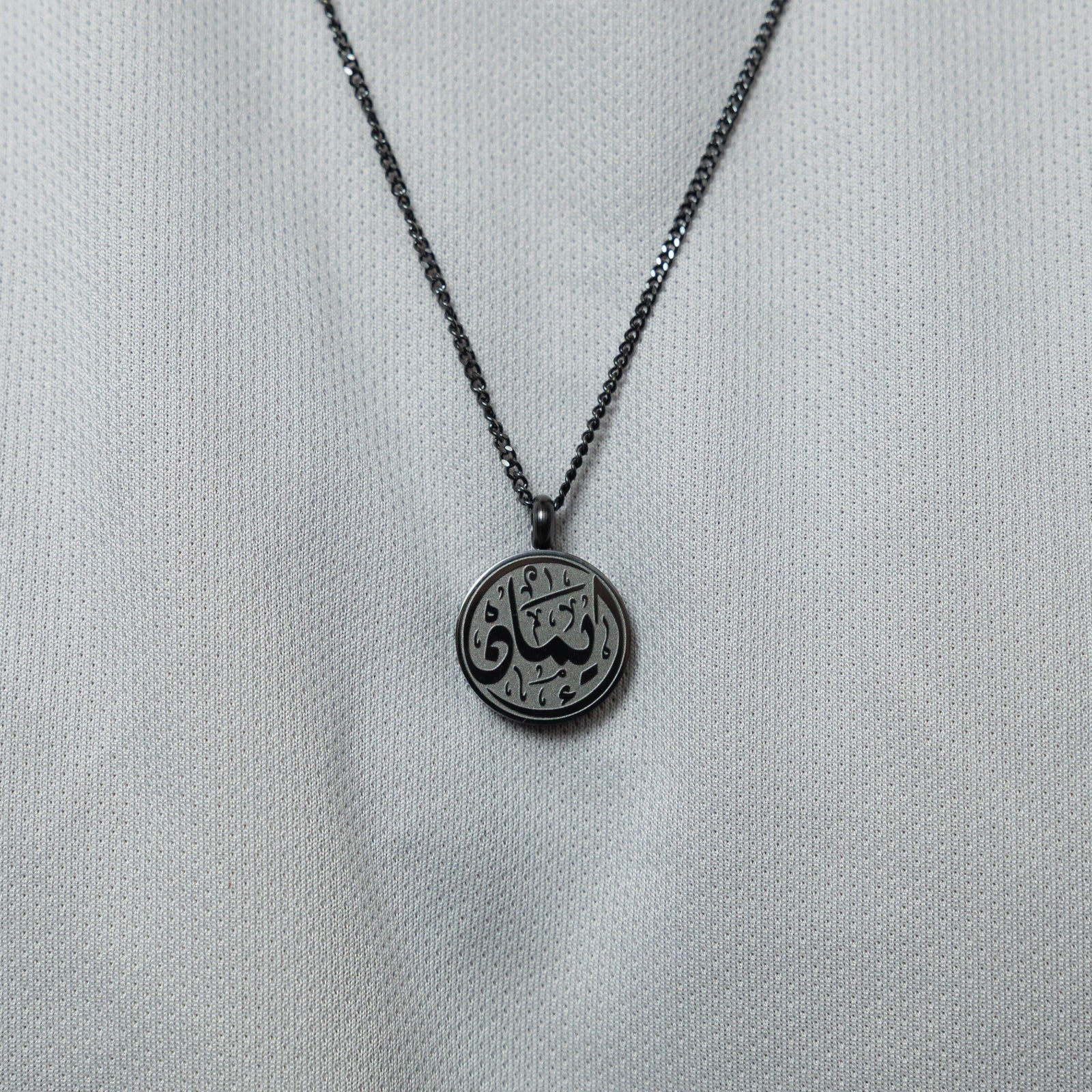 A close-up of the black Faith Token pendant on a  white textured shirt, showing the engraved Arabic script "Iman".