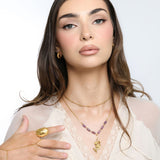 Woman wearing an 18k gold plated layered necklace with a flower stem pendant and purple tube beads, paired with a matching gold ring and hand chain