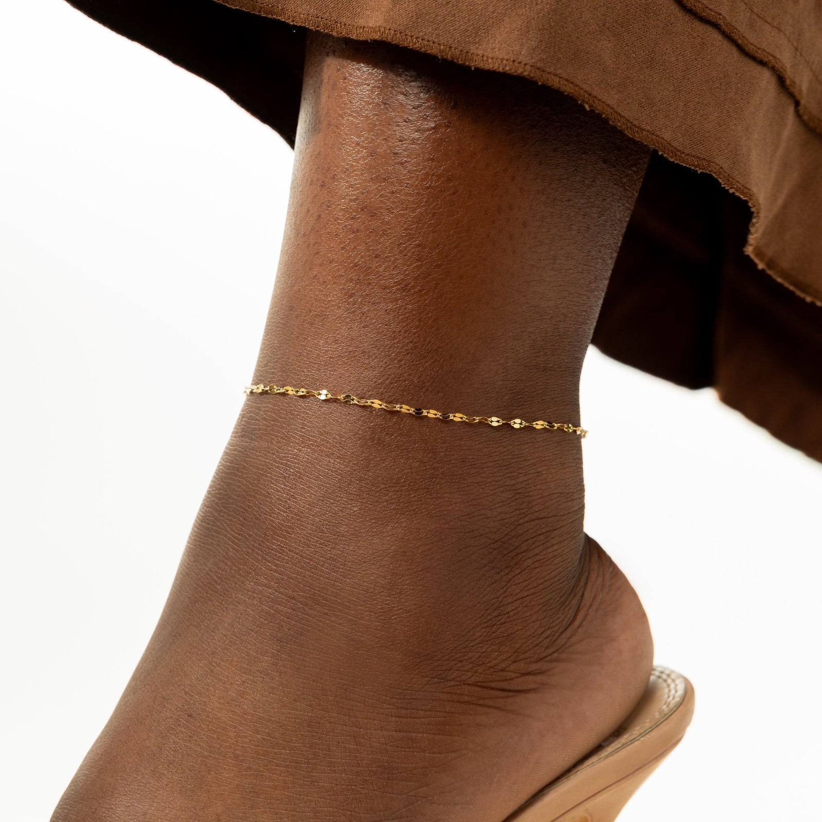 Close-up of a gold flutter chain anklet worn above the ankle, styled with a brown pants and open sandals. 