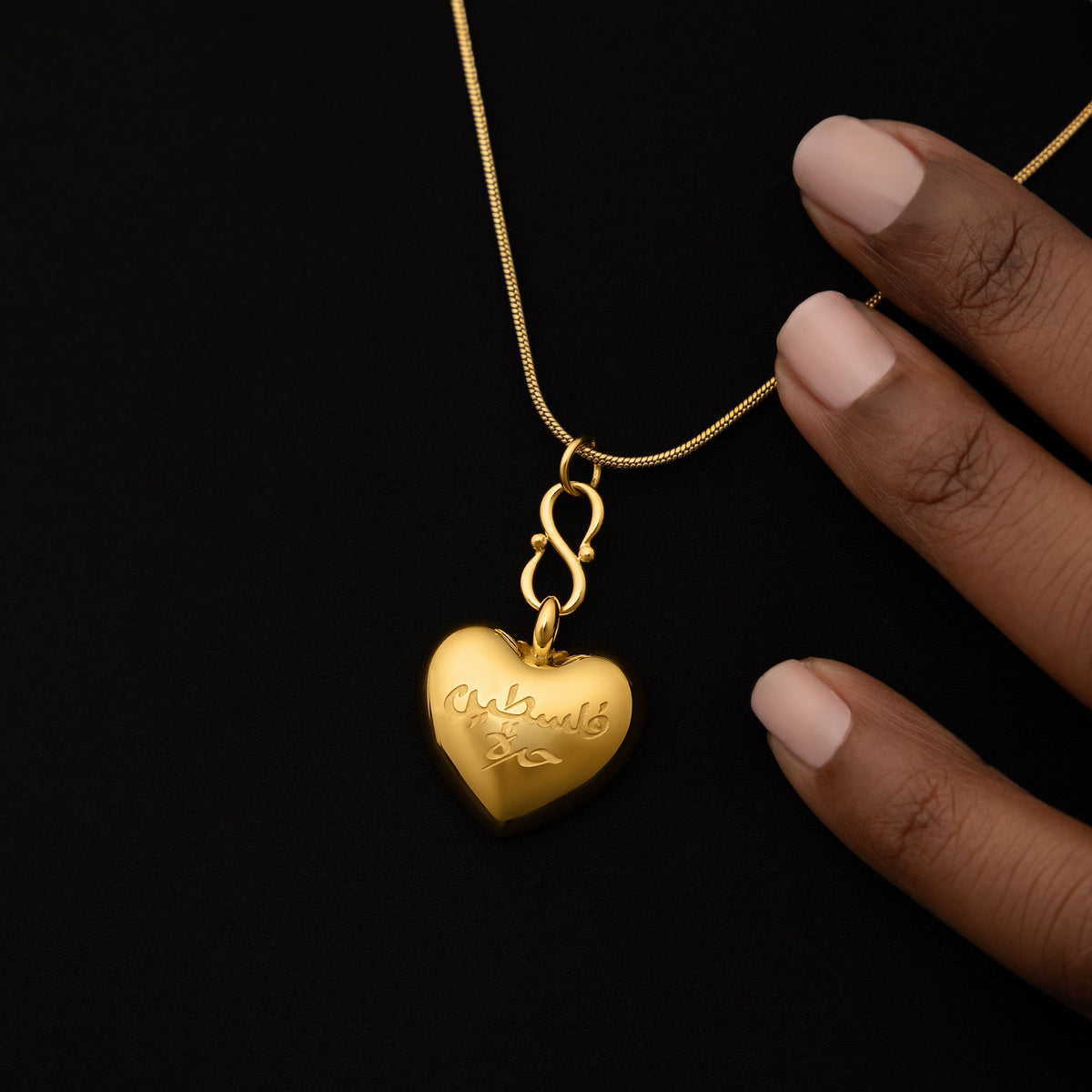 Close up of the gold "Free Palestine" Heart Necklace resting against a black background. The model is touching the thin gold chain of the necklace.