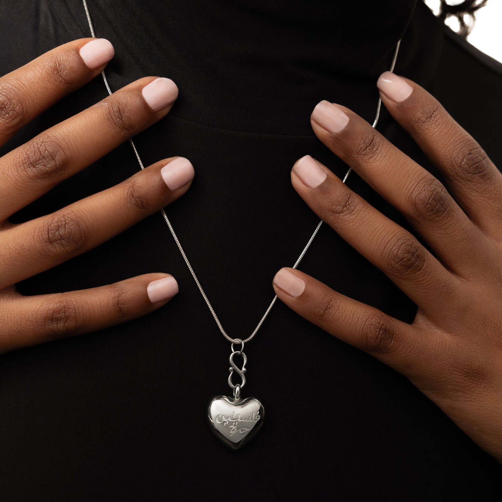 Close up of the silver "Free Palestine" Heart Necklace. The model is touching the thin silver chain with their fingers.