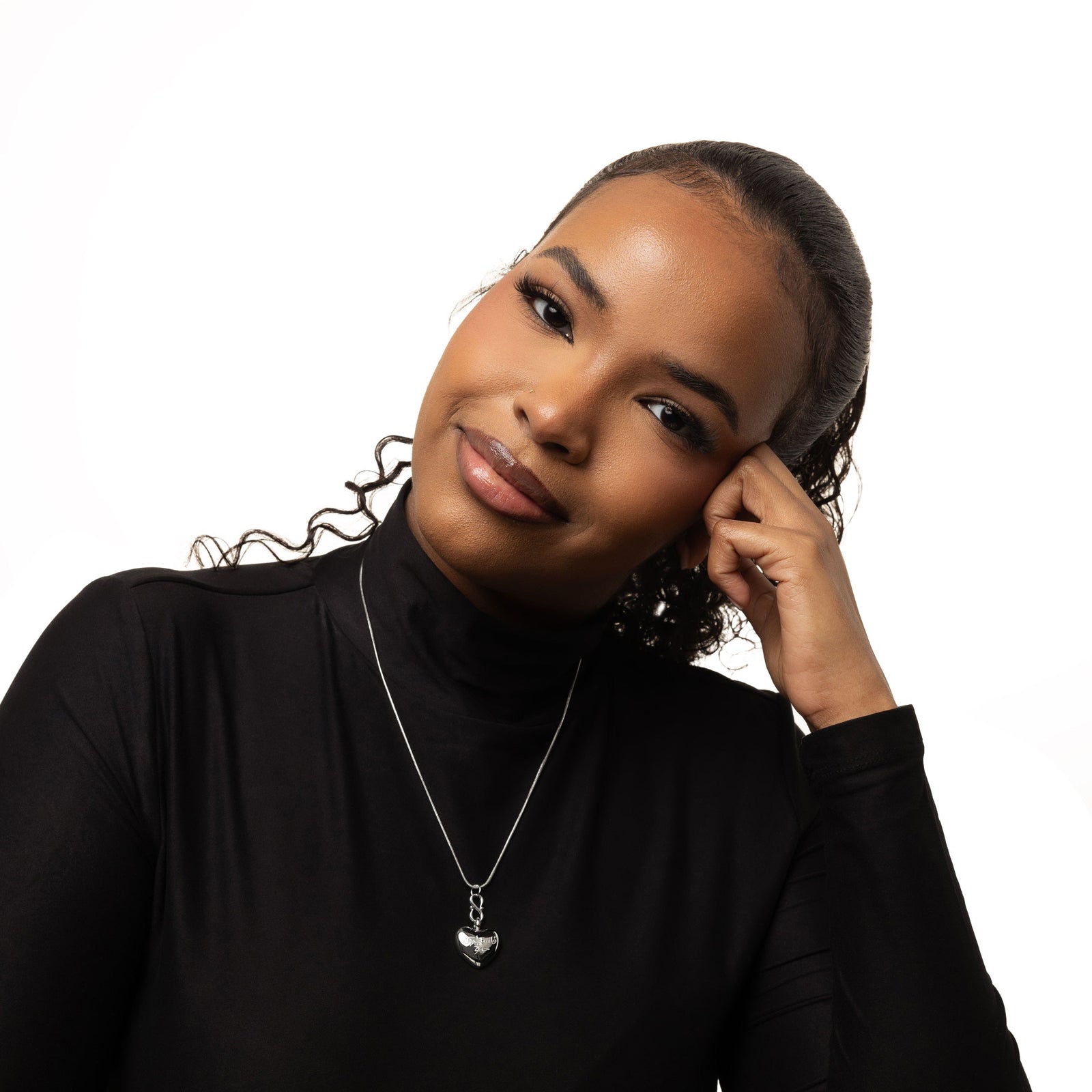 Photo of the model wearing a fitted black long-sleeve top with a high neckline. Around her neck is a delicate chain necklace with a small, heart-shaped pendant.