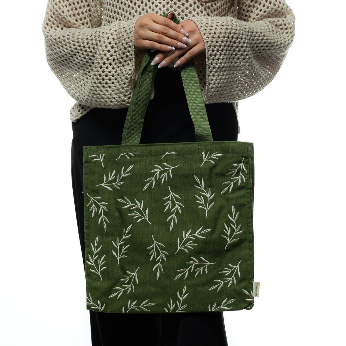 Woman wearing a beige knit sweater holding a forest green tote bag with embroidered olive branch motifs.