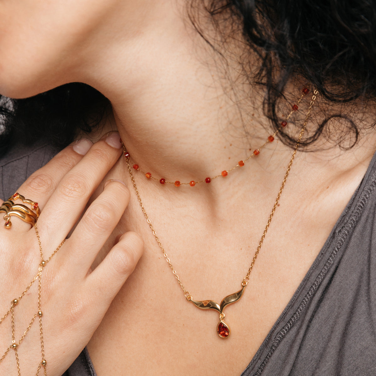 Model styled in 18k gold-plated Nominal jewelry featuring the Essential Red Agate Micro Chain layered with the Garnet Stone Keffiyeh Necklace, paired with the Keffiyeh Droplet Ring Stack and red beaded hand chain.