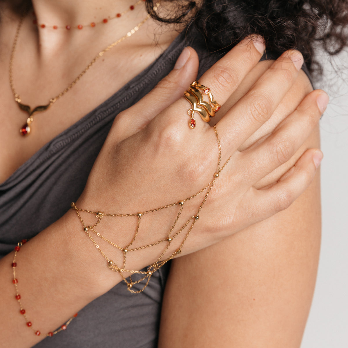 Model styled in 18k gold-plated Nominal jewelry featuring the Garnet Stone Weaving Keffiyeh Ring paired with the Red Bead Fishnet Hand Chain, Garnet Stone Keffiyeh Necklace, and Essential Red Agate Micro Chain.