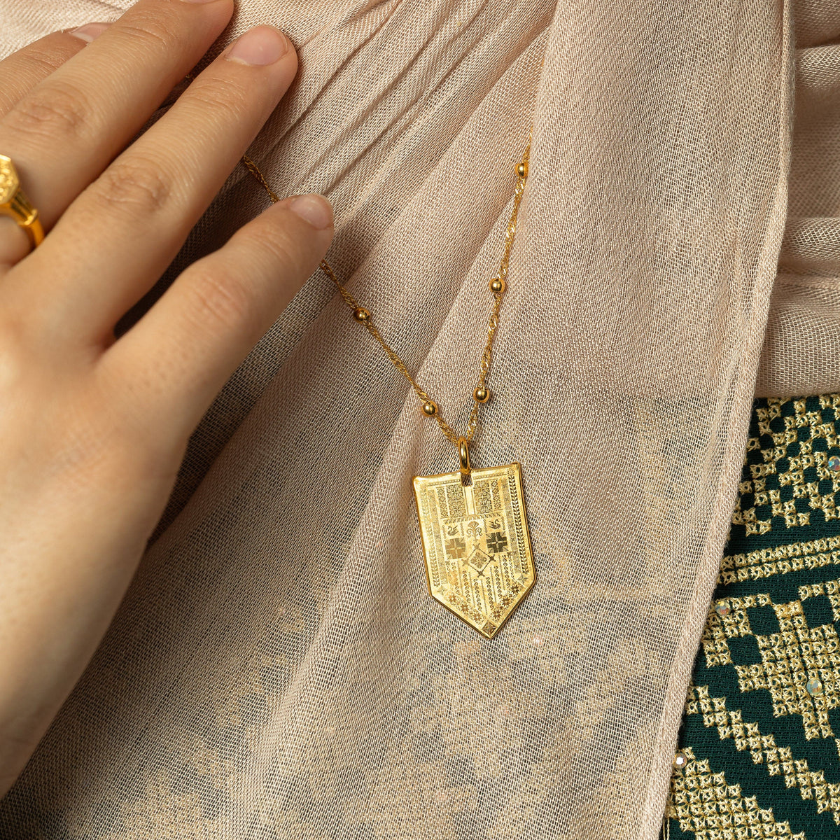 In the photo, a model touching the dotted gold chain of the Hanging Tatreez Necklace showing the design of the pointed tag pendant.