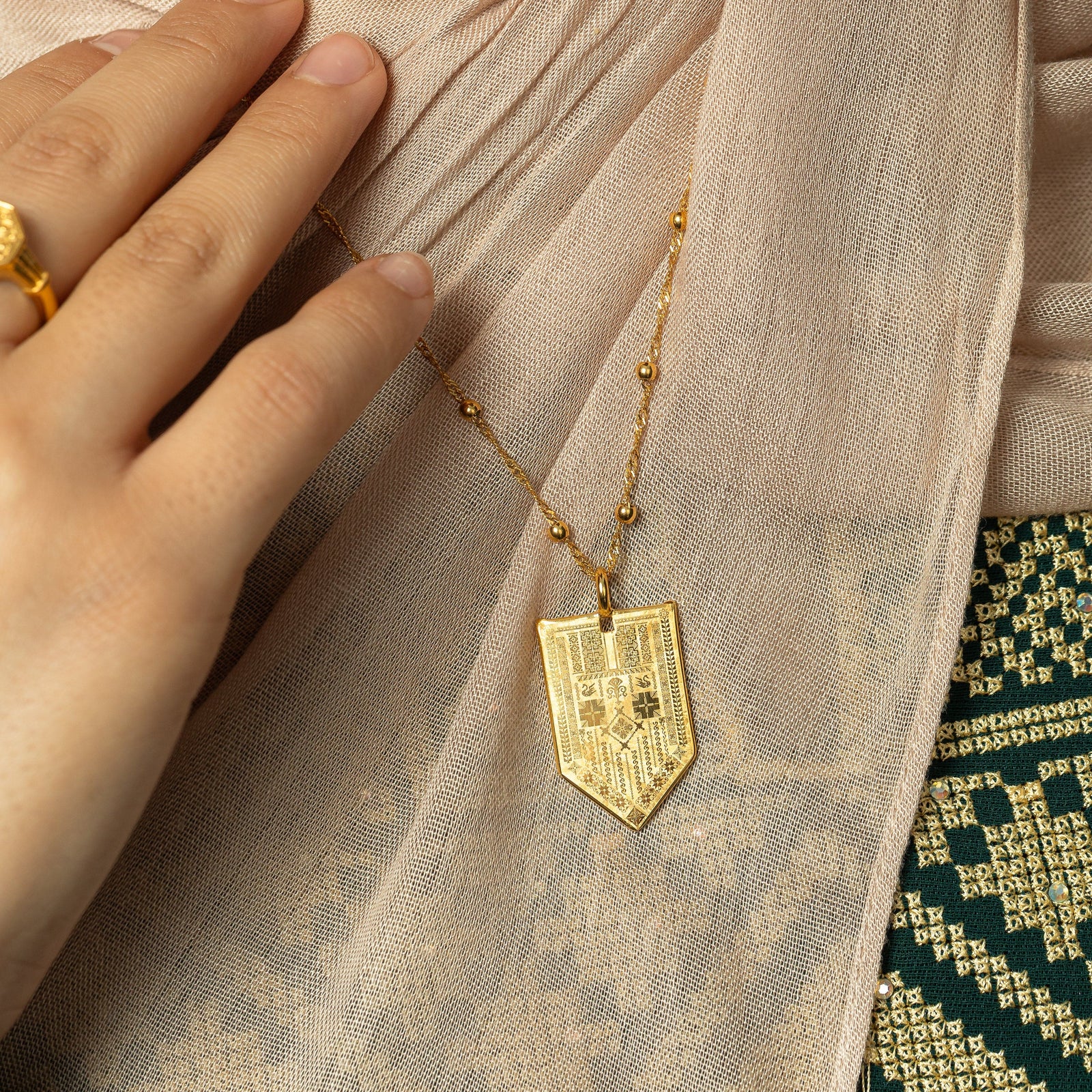 In the photo, a model touching the dotted gold chain of the Hanging Tatreez Necklace showing the design of the pointed tag pendant.
