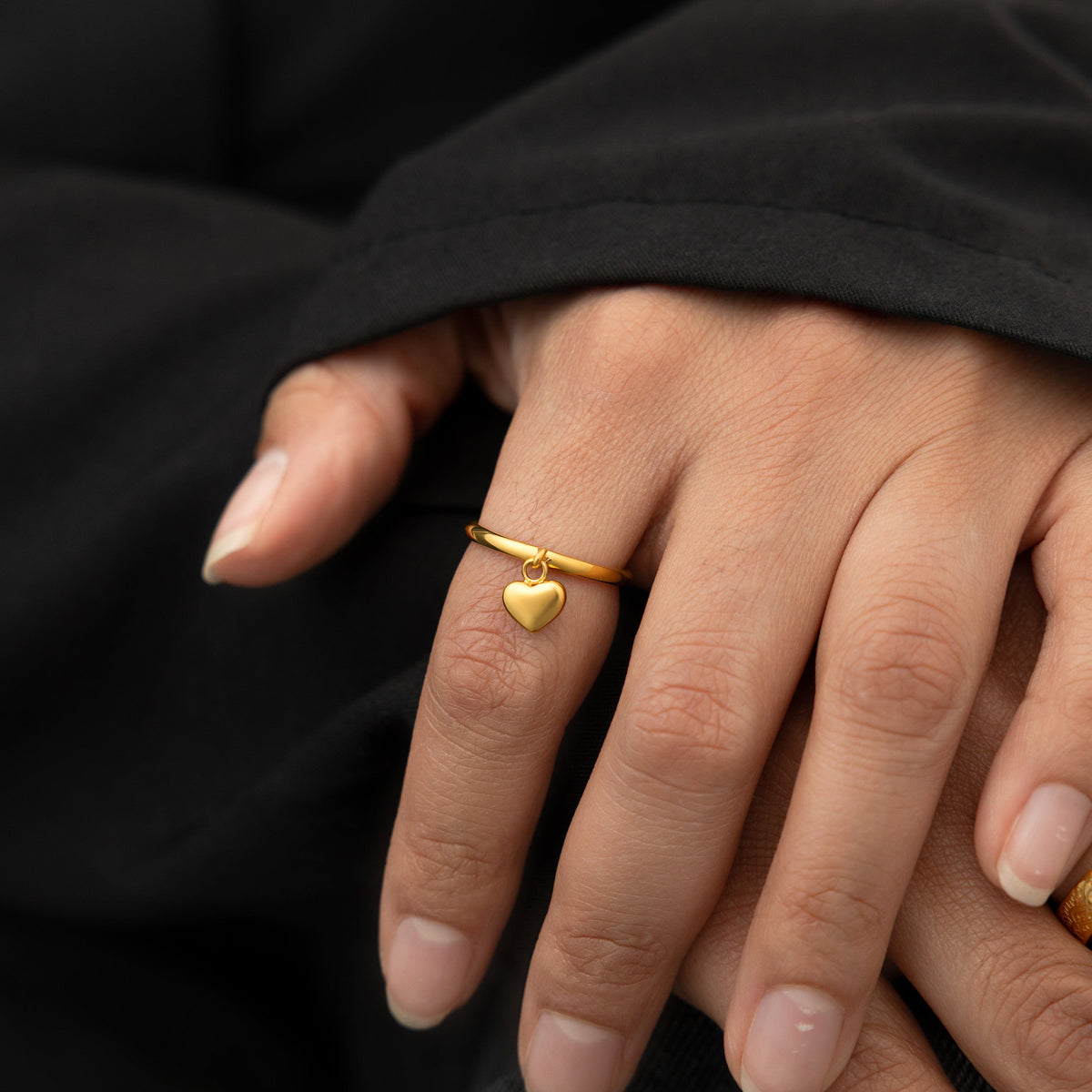 Hand wearing a slim gold ring with a dangling heart charm, styled against a black sleeve.